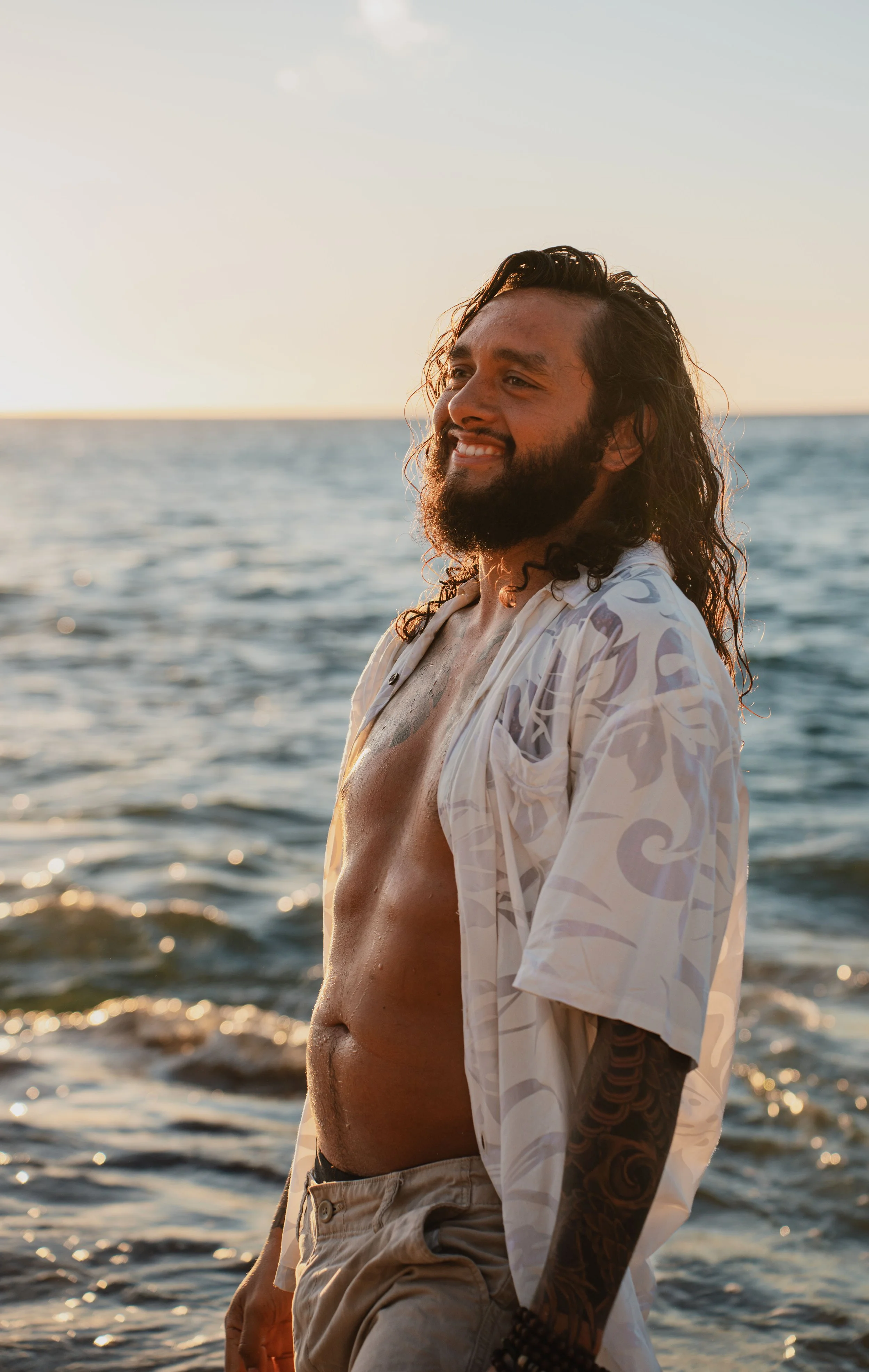 Male smiling during a photoshoot while in the water of a beach in Oahu, Hawaii wearing a unbutton shirt and shorts. 