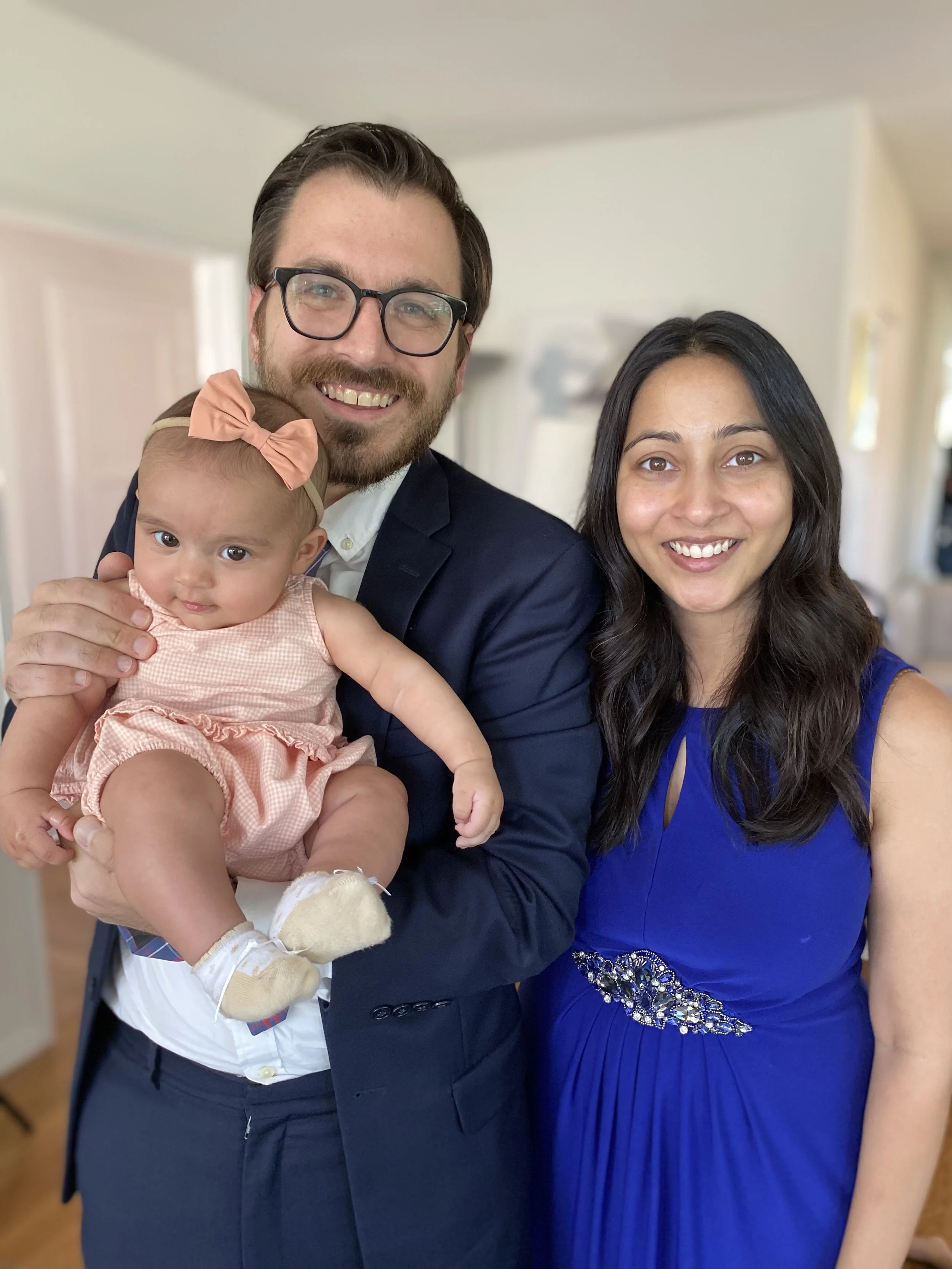 A smiling man in glasses, a woman in a blue dress, and a baby girl with a peach bow headband pose together indoors.