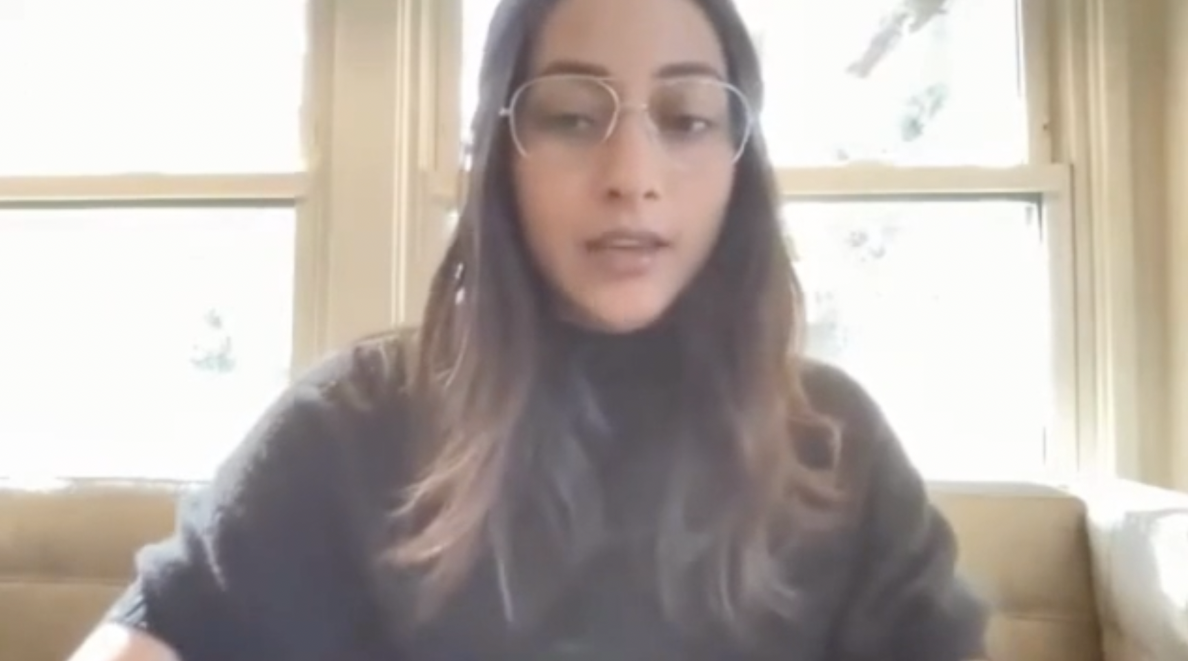 A young woman with long brown hair, wearing glasses and a black turtleneck, sitting on a beige couch in front of large windows with white frames, during a video call or virtual meeting.