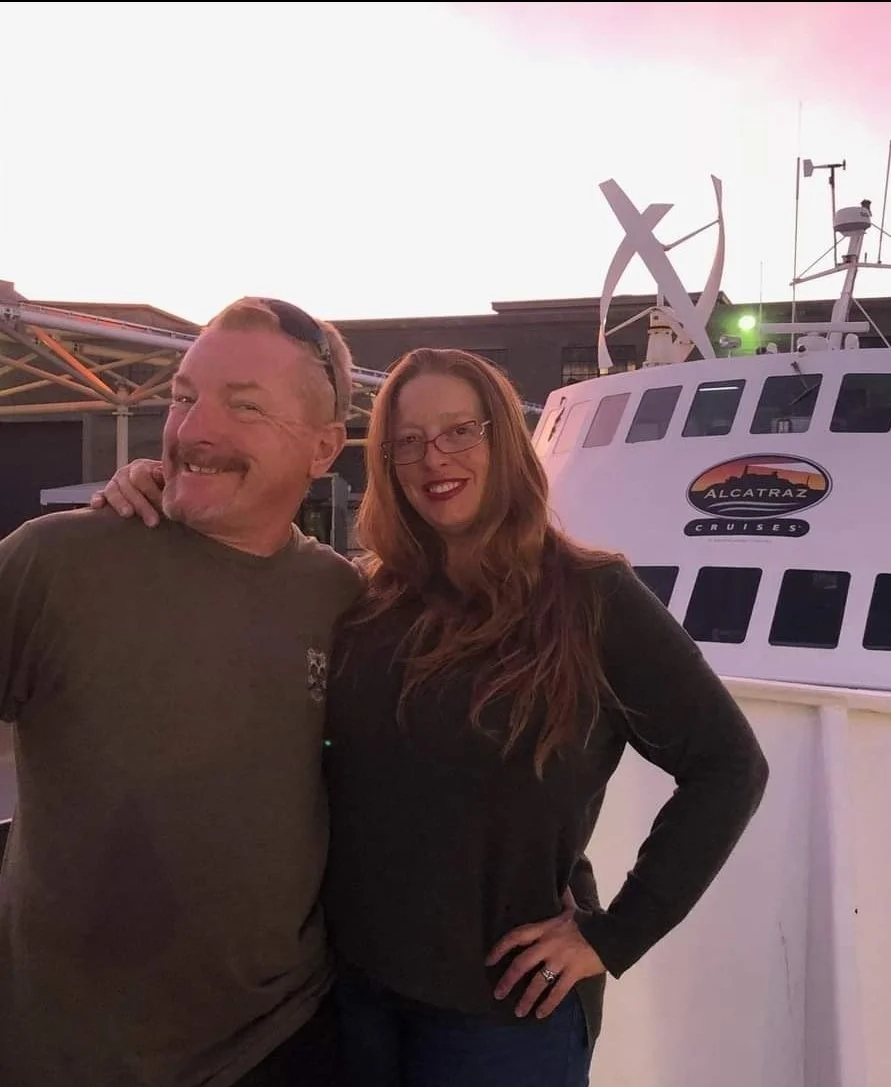 Smiling owners of the Niles Flying A, Rene and Krysten, standing together outdoors at sunset, with a boat in the background, which has a logo reading 'Alcatraz Cruises.'