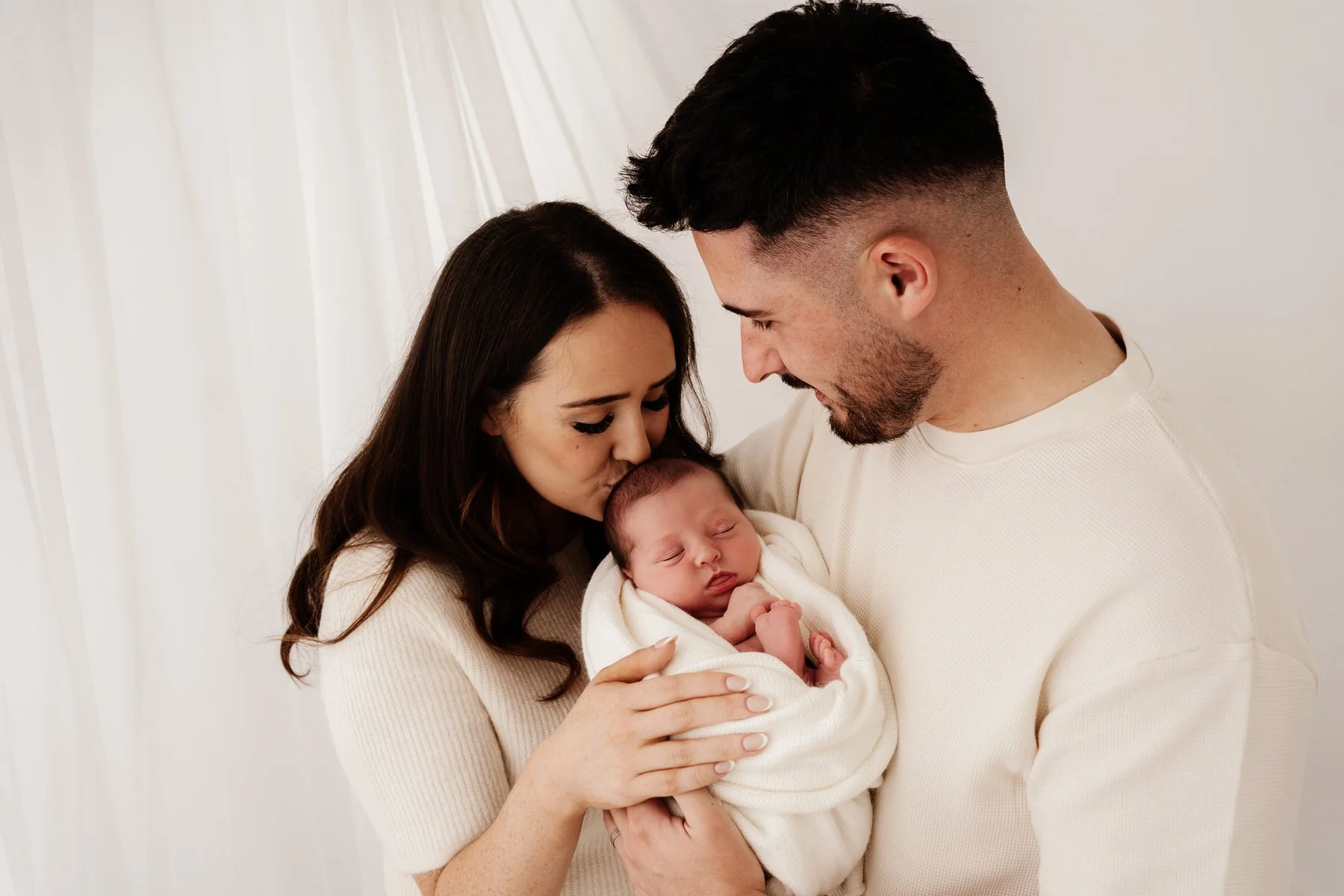 Dad holding  his newborn baby while mum kisses her head.