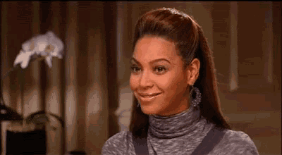 A woman with long hair styled in a high ponytail, wearing a grey turtleneck top, smiling in an indoor setting.