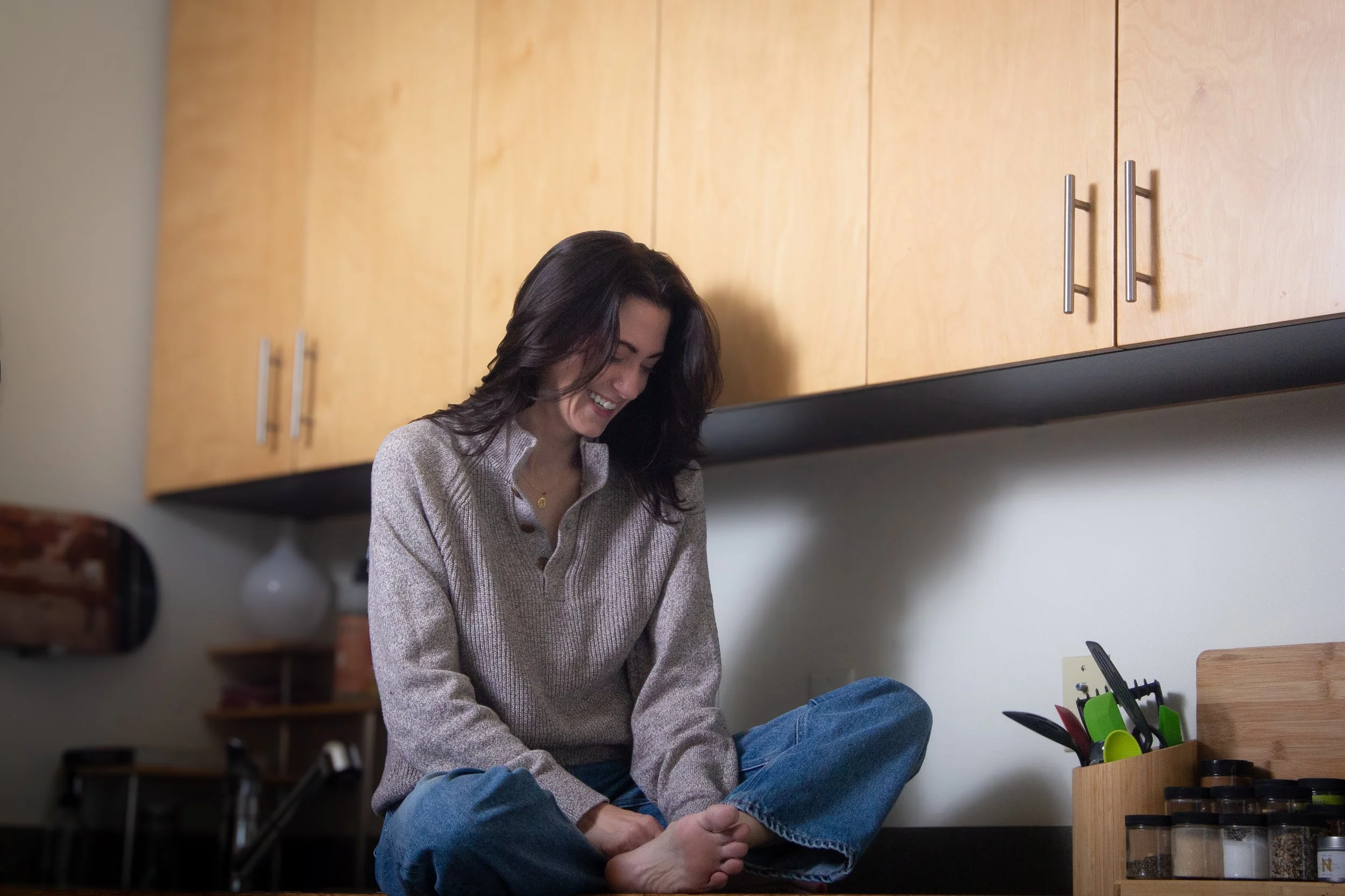 A woman who can help make your every day life easier. She has dark hair and a light-colored sweater, and is sitting cross-legged on her kitchen counter. Feeling at ease in her own home, she smiling and appears to be fully relaxed.