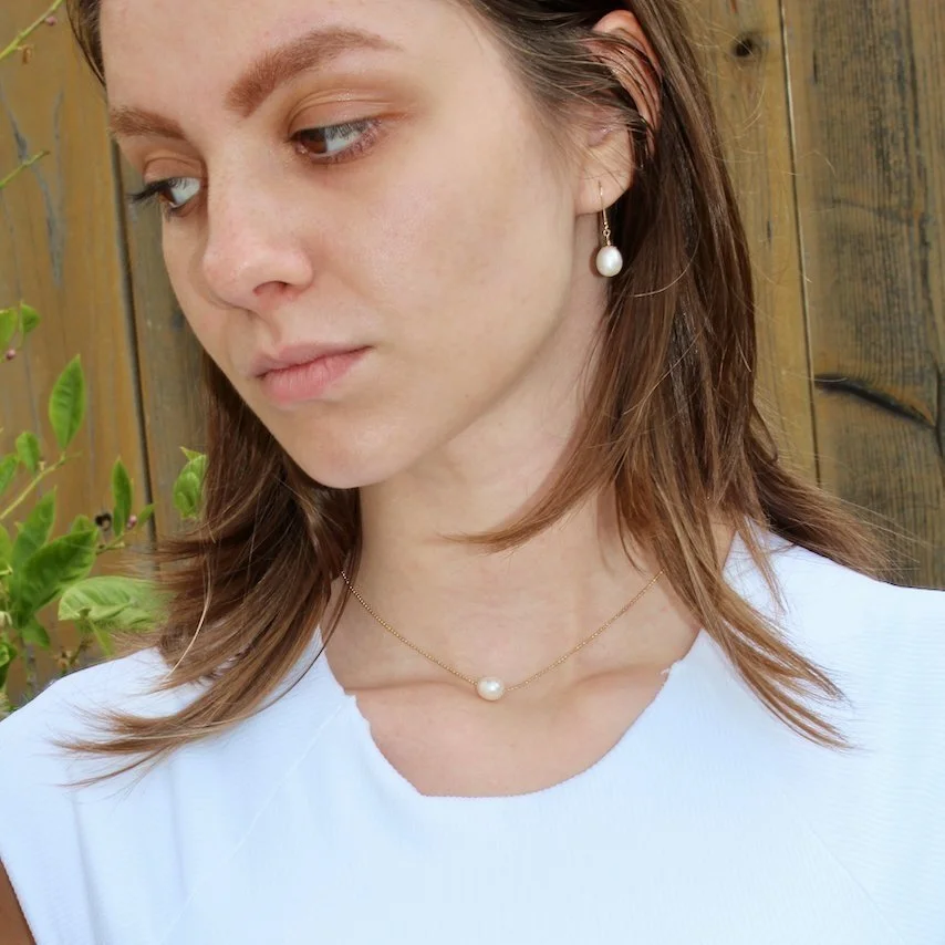 A woman with brown hair and fair skin wearing pearl jewelry and a white top, standing outdoors near a wooden fence and greenery.