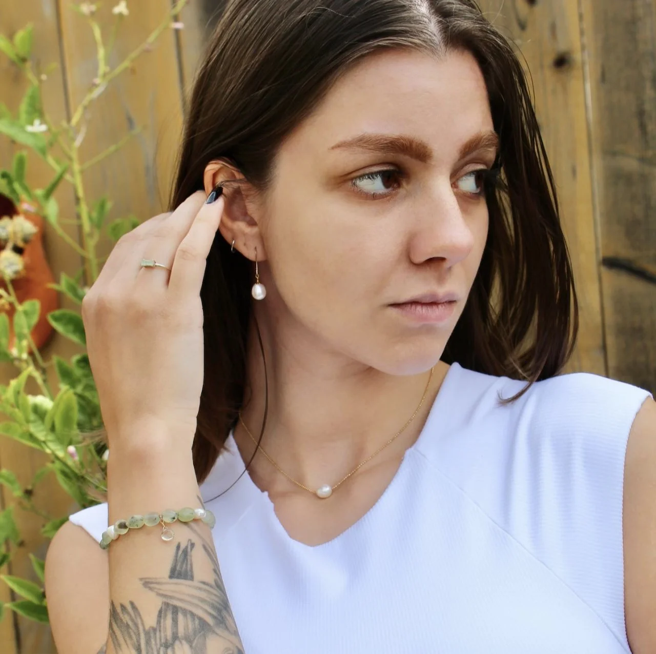 Close-up of a young woman with dark brown hair, wearing pearl and gold jewelry, adjusting her earring outdoors with a wooden fence and green plants in the background.