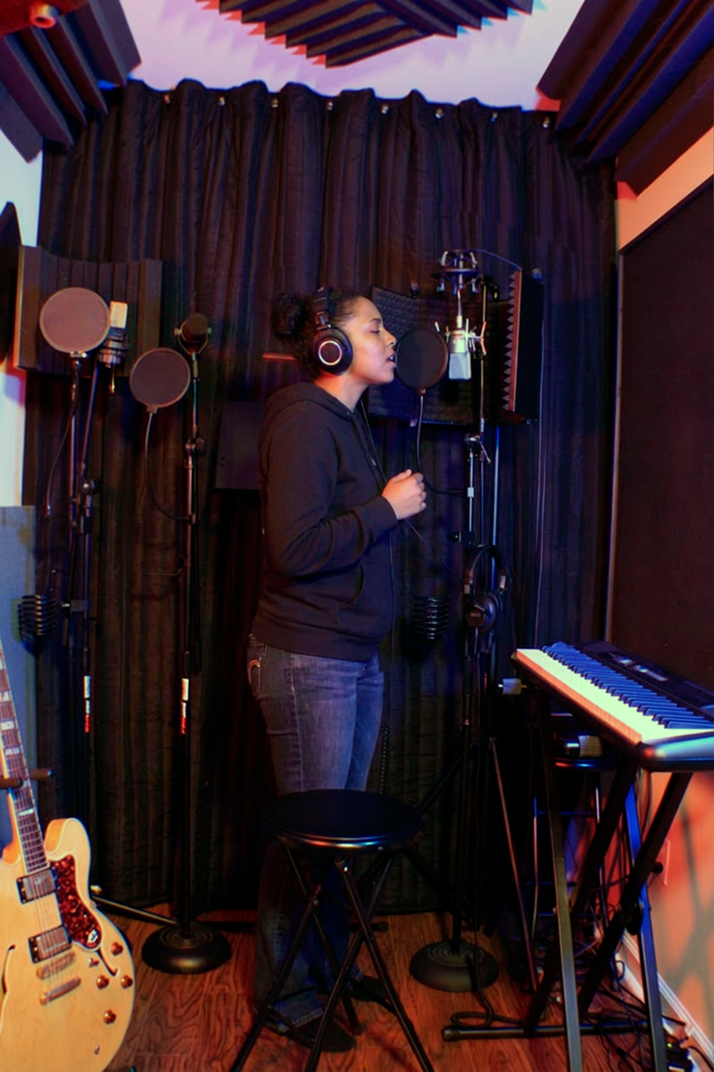 A woman with headphones singing into a microphone in a home recording studio, with a keyboard and guitar in the room.