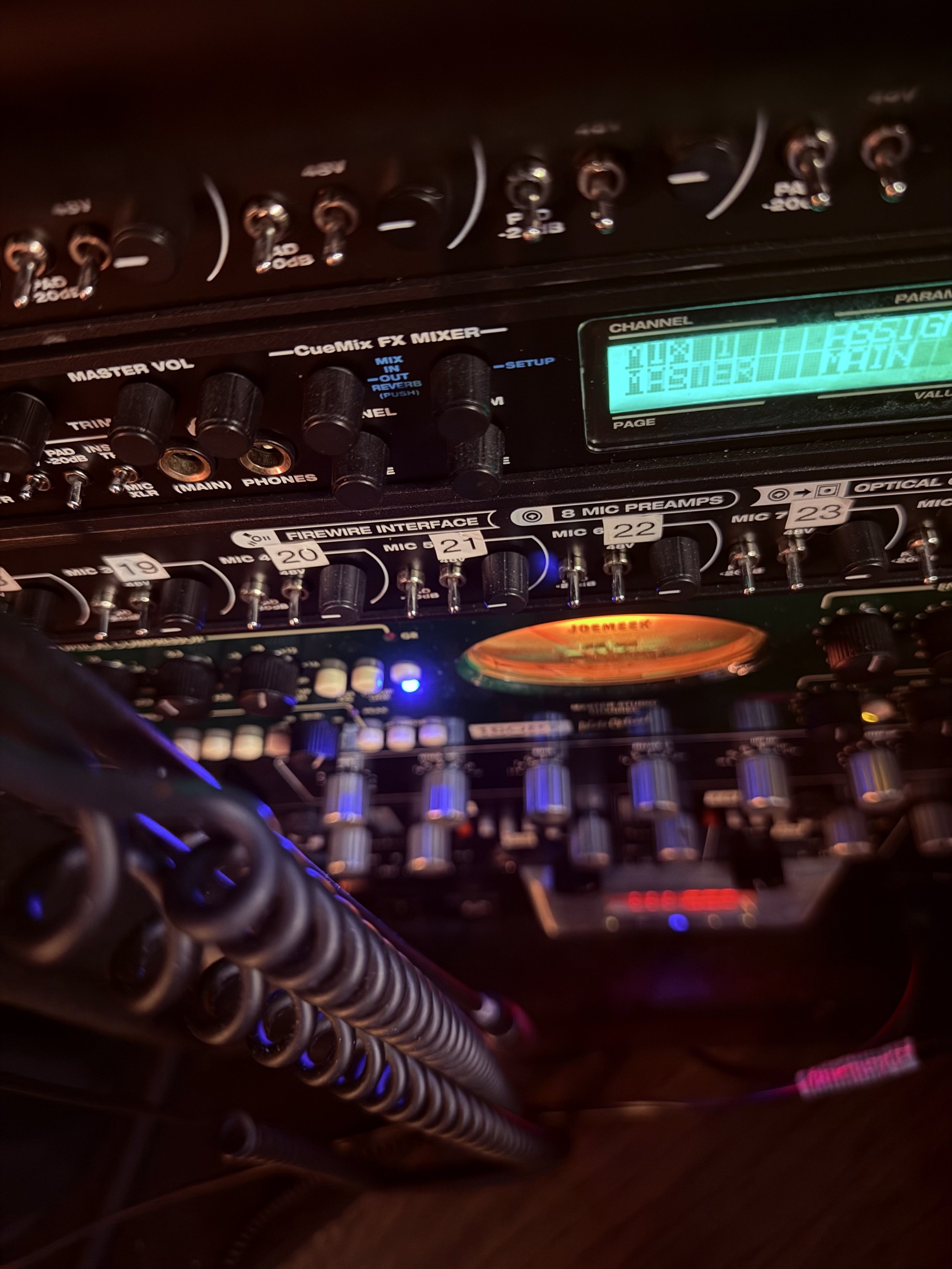 Close-up of a professional audio mixing console with various controls, knobs, and a digital display illuminated in teal, showing settings and levels.