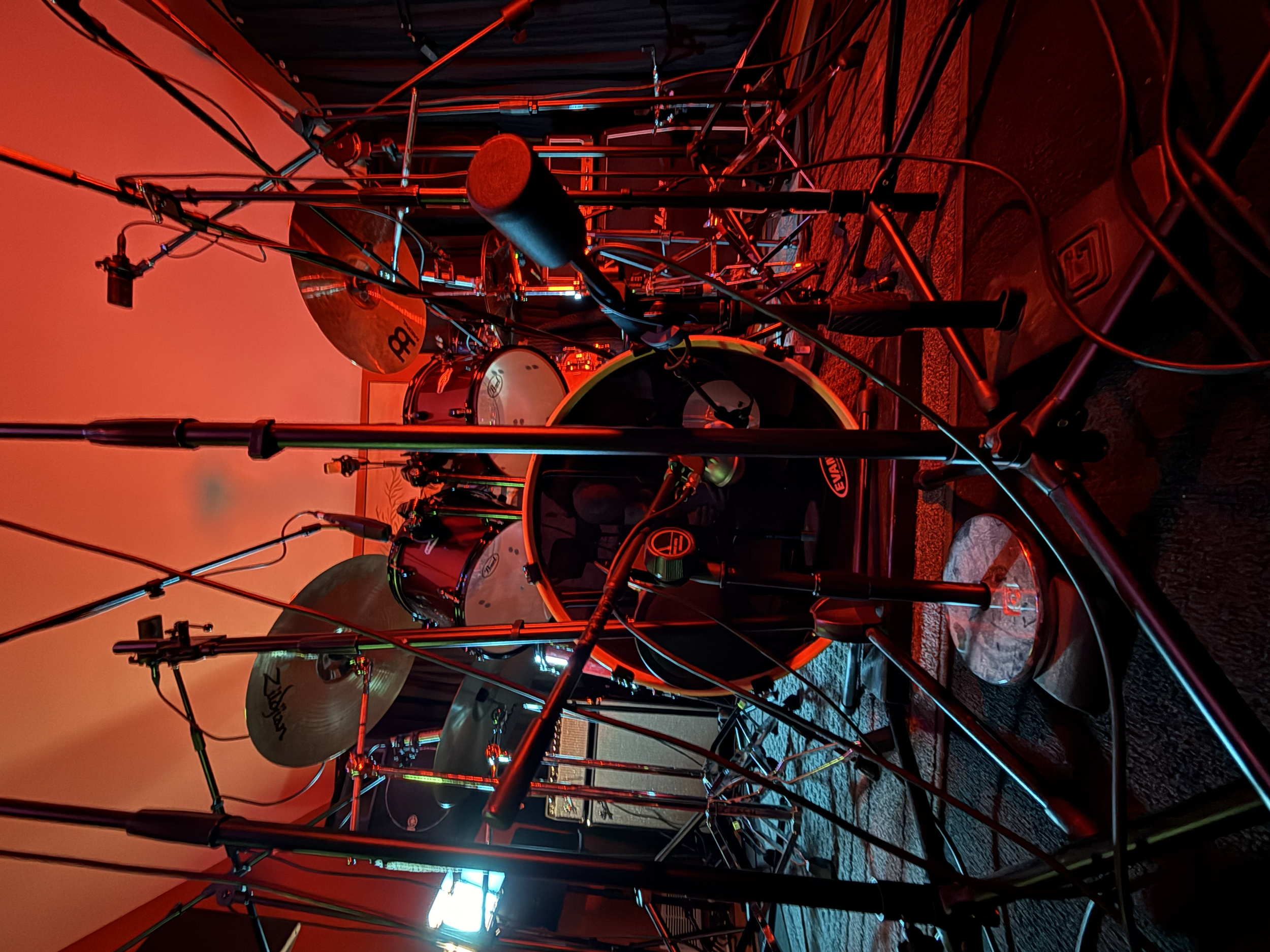 A rooftop drum set illuminated with blue and red lights, including cymbals, drums, microphone, and stands, with a sunset sky in the background.