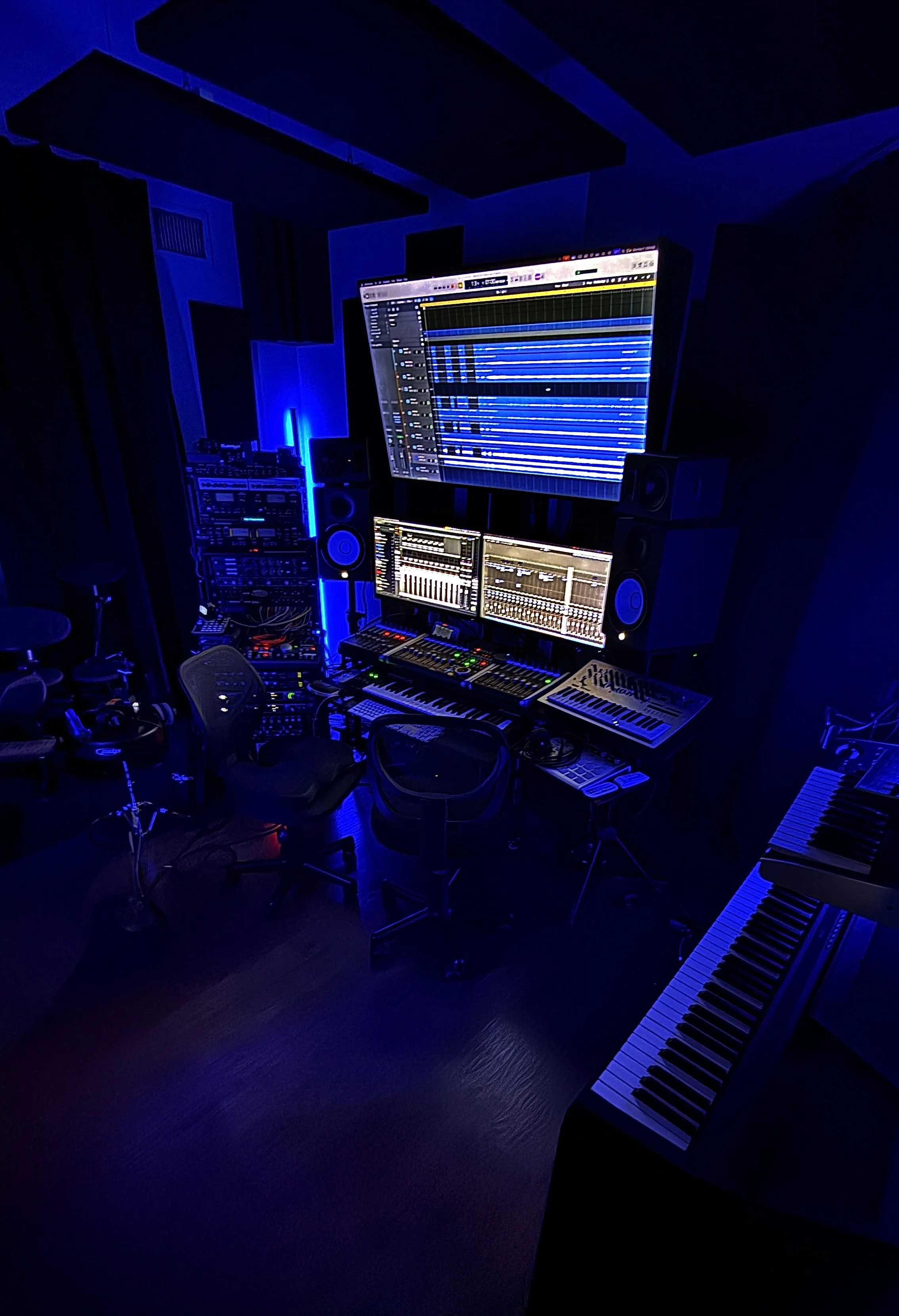 Music studio with computer monitors, audio equipment, keyboard instruments, and blue lighting.