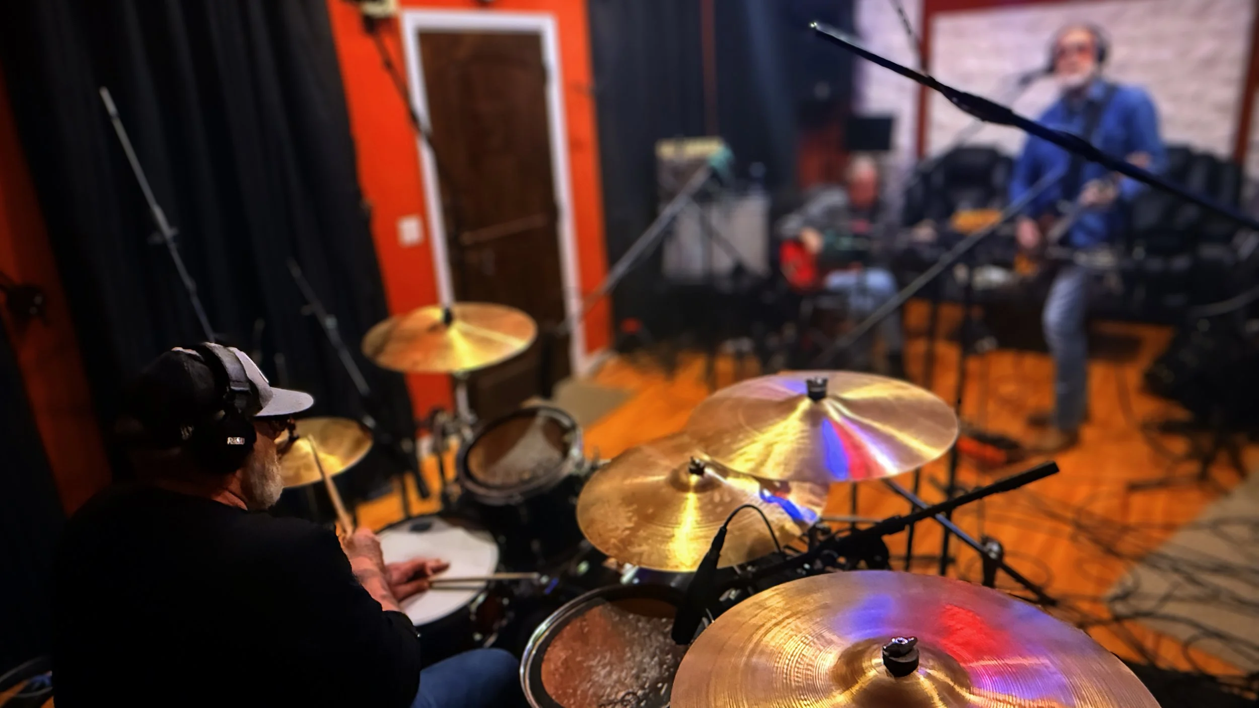 A drummer wearing headphones and a cap playing drums in a recording studio, with a singer and another musician in the background.