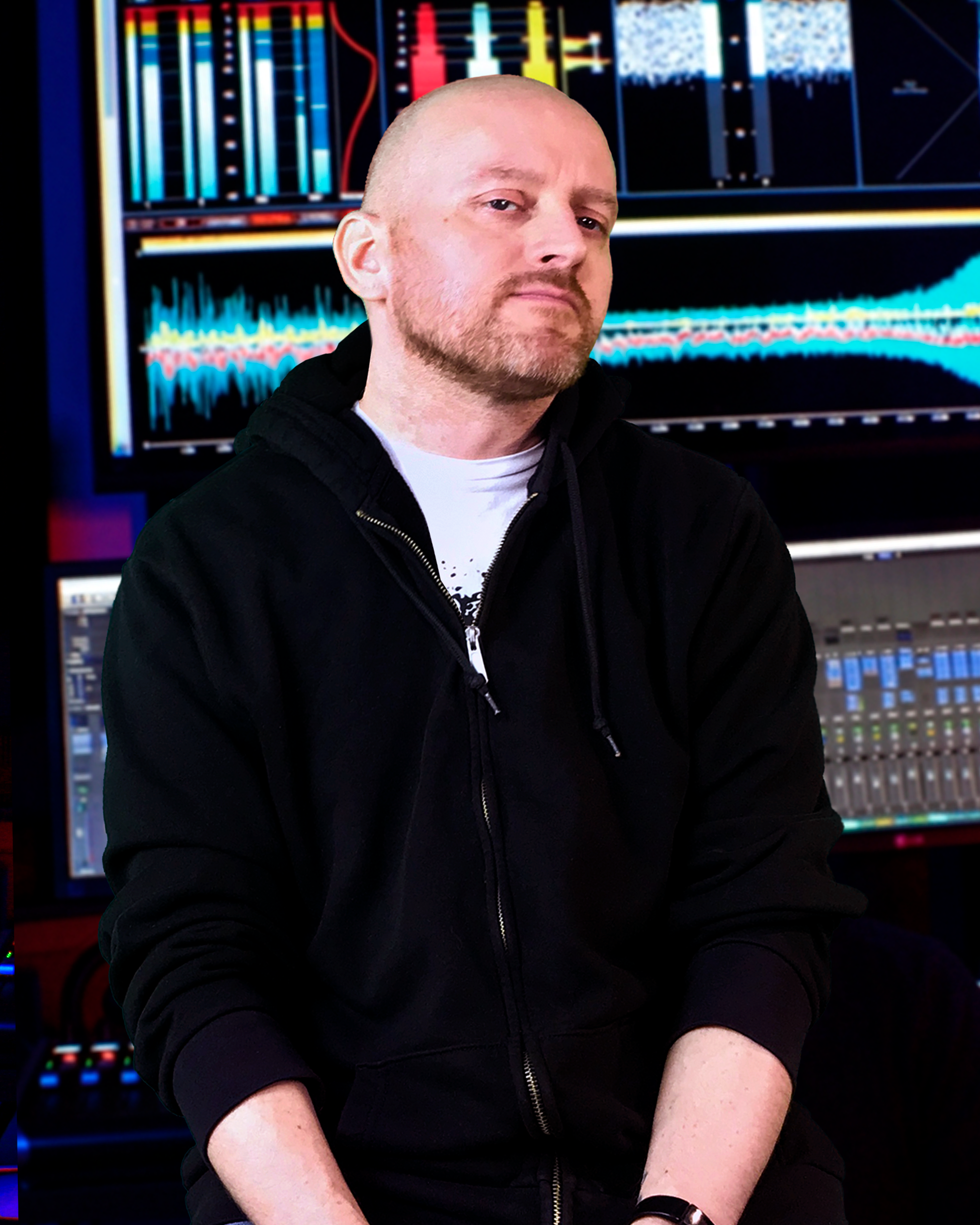 Man with a blond beard wearing a black hoodie sitting in front of a digital audio workstation with colorful audio waveforms and mixing panels on screens.