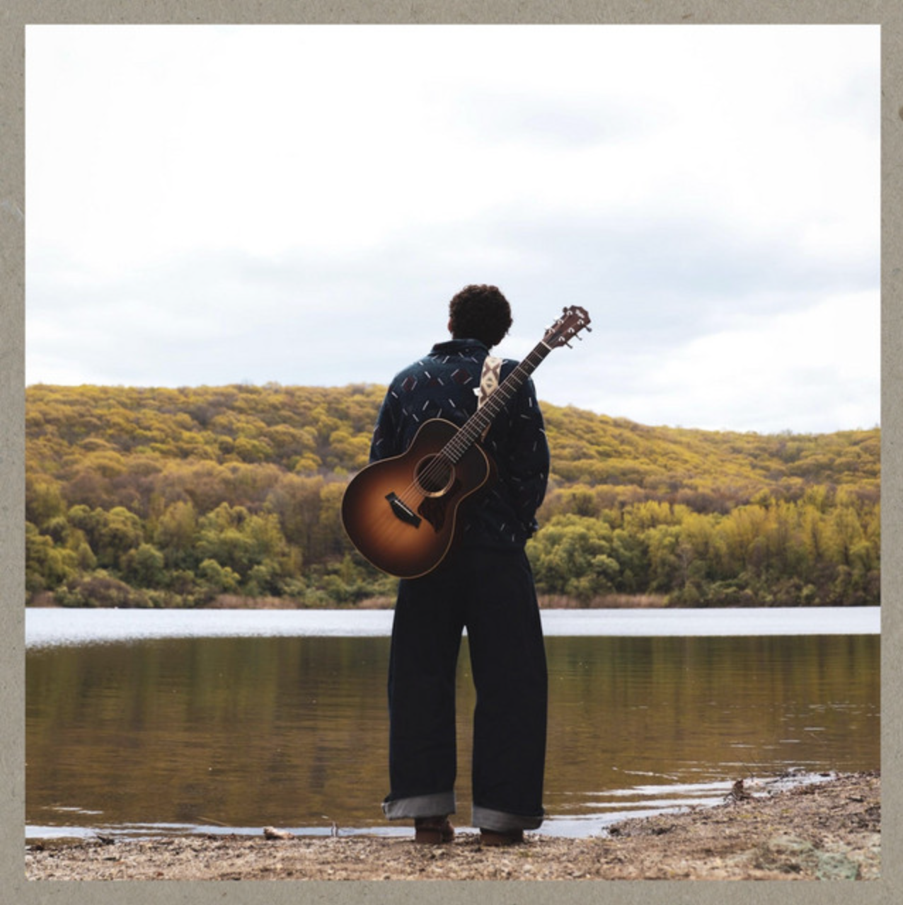 Person standing by a lake with a guitar on their back, overlooking a wooded landscape with yellow and green trees under cloudy sky.