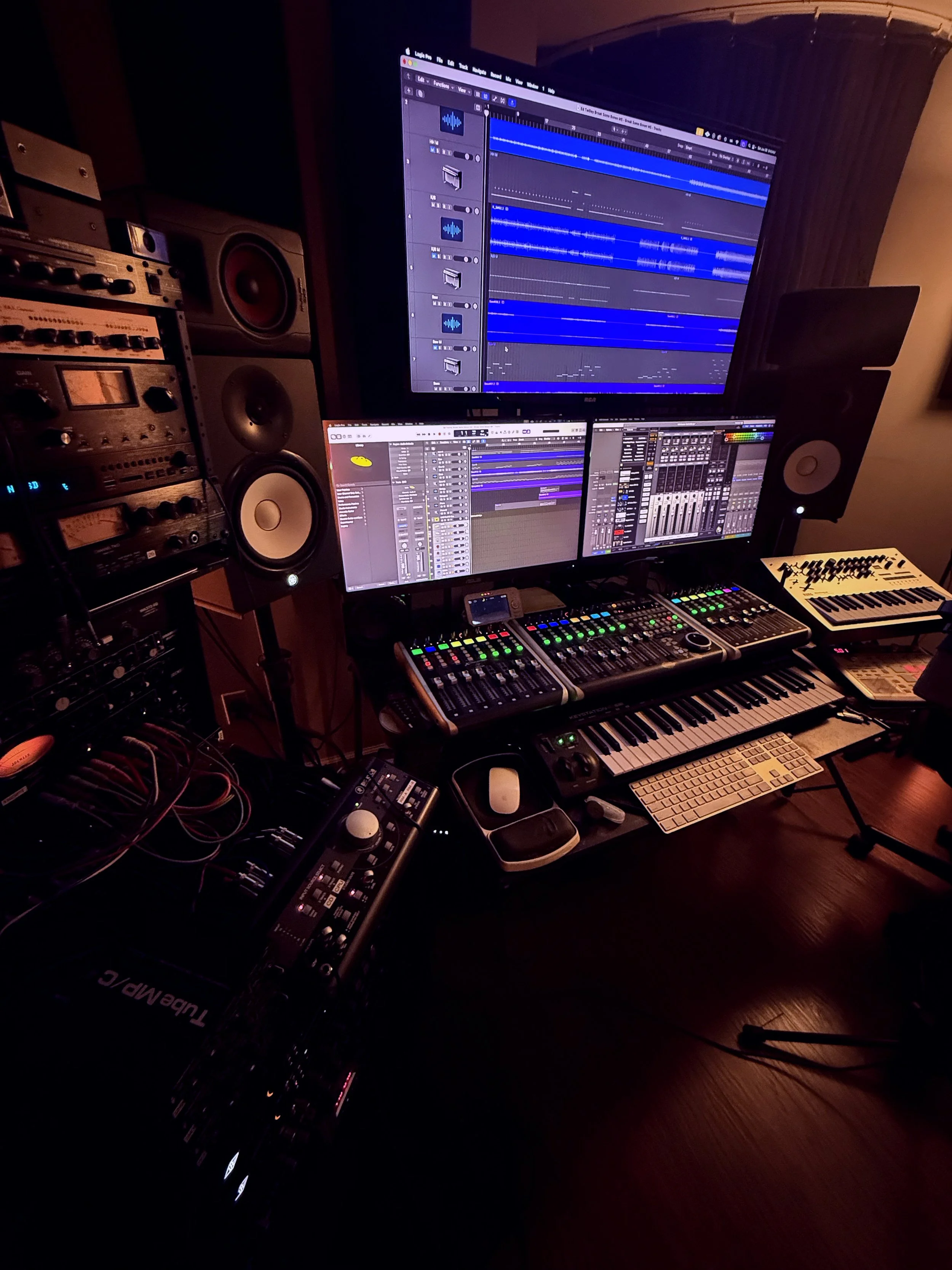 Music production studio with multiple computer monitors displaying digital audio workstations, audio mixing consoles, studio speakers, keyboards, and audio equipment.