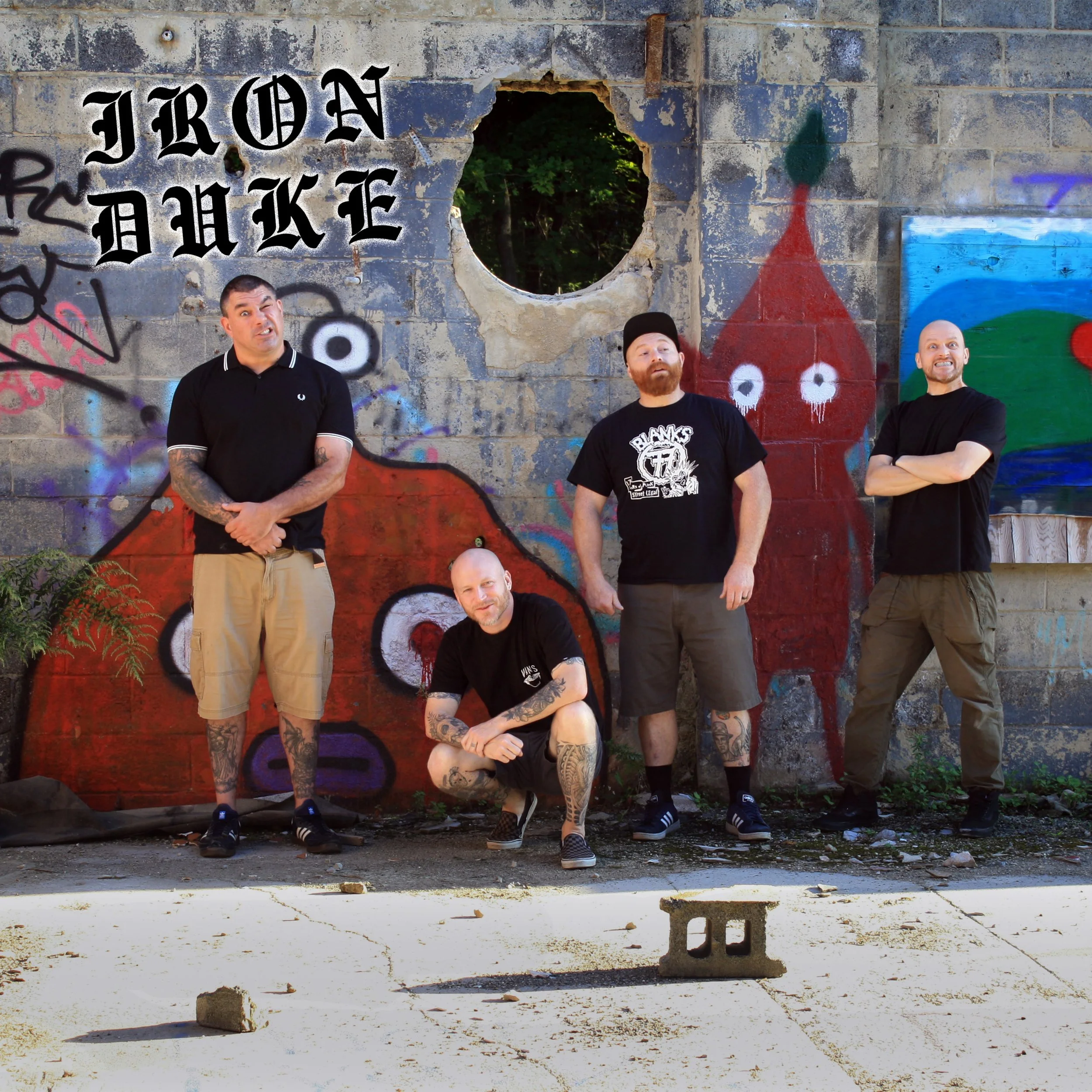 Four men with tattoos and casual clothing posing in front of a graffiti-covered brick wall, with a hole in the wall showing trees behind.