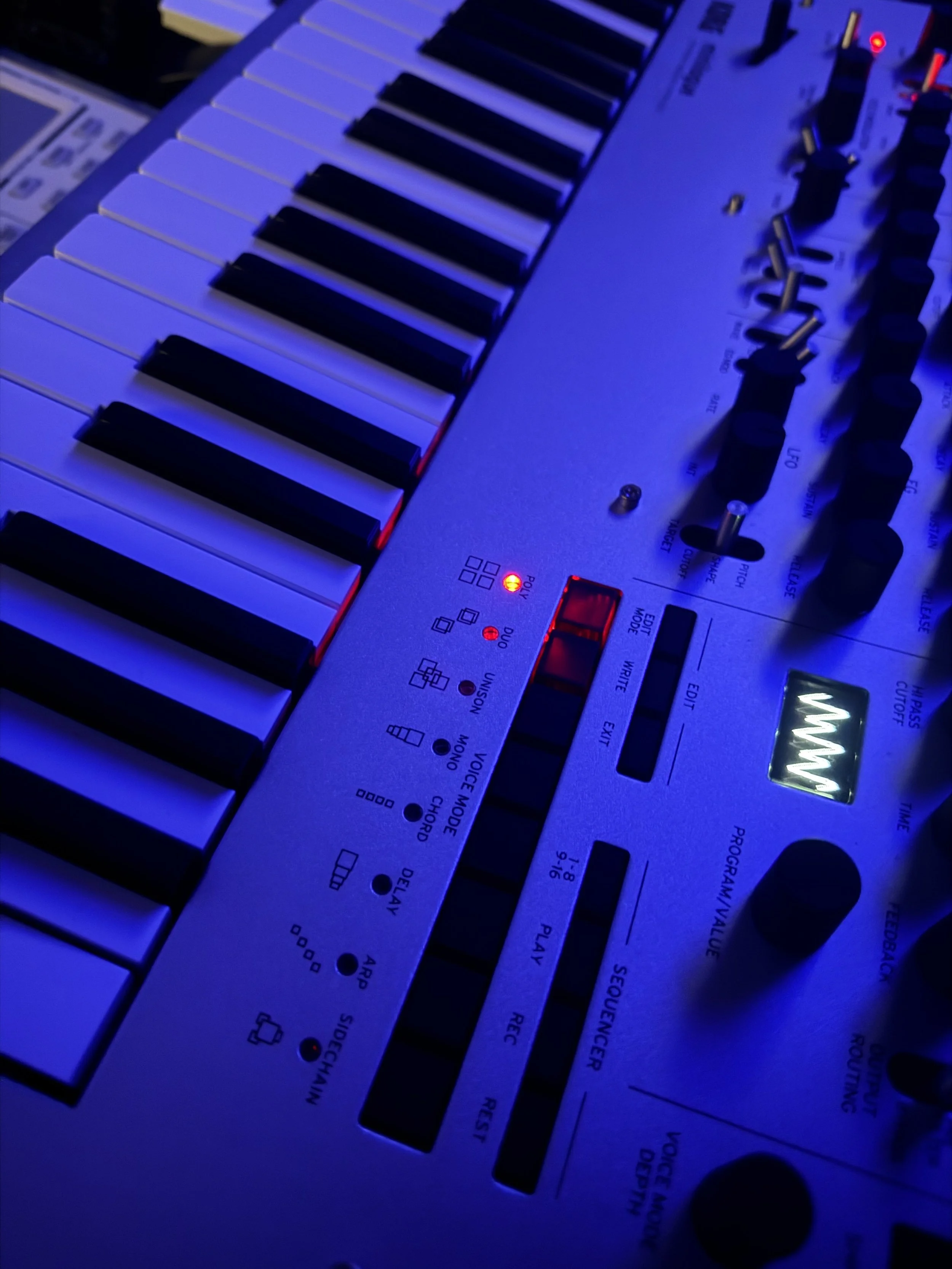 A synthesizer keyboard with black and white keys, illuminated by blue and purple lighting, featuring various control knobs and buttons.