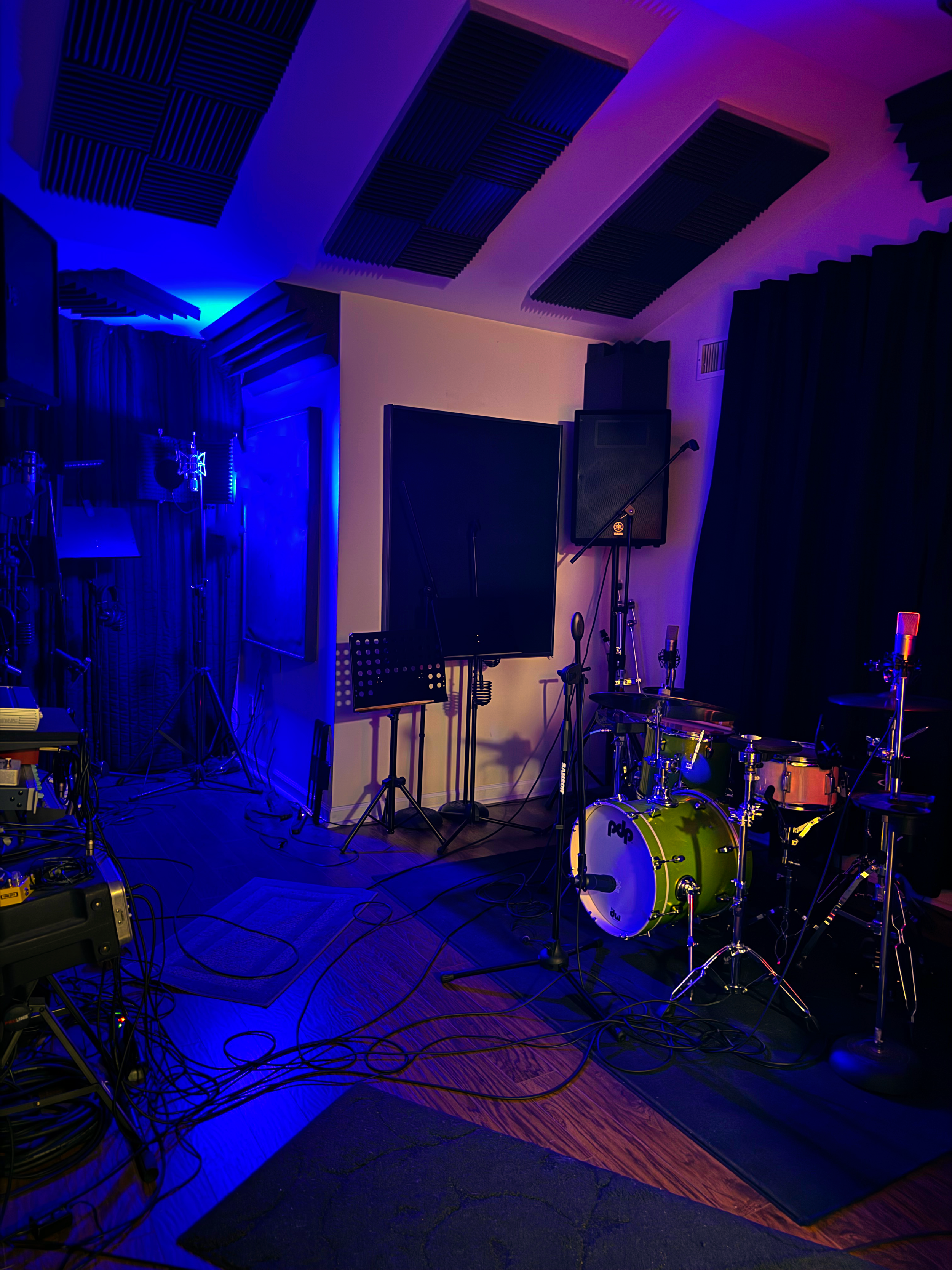 Music recording studio with drum set, microphone, speakers, and soundproofing panels on walls, dimly lit with blue lighting.