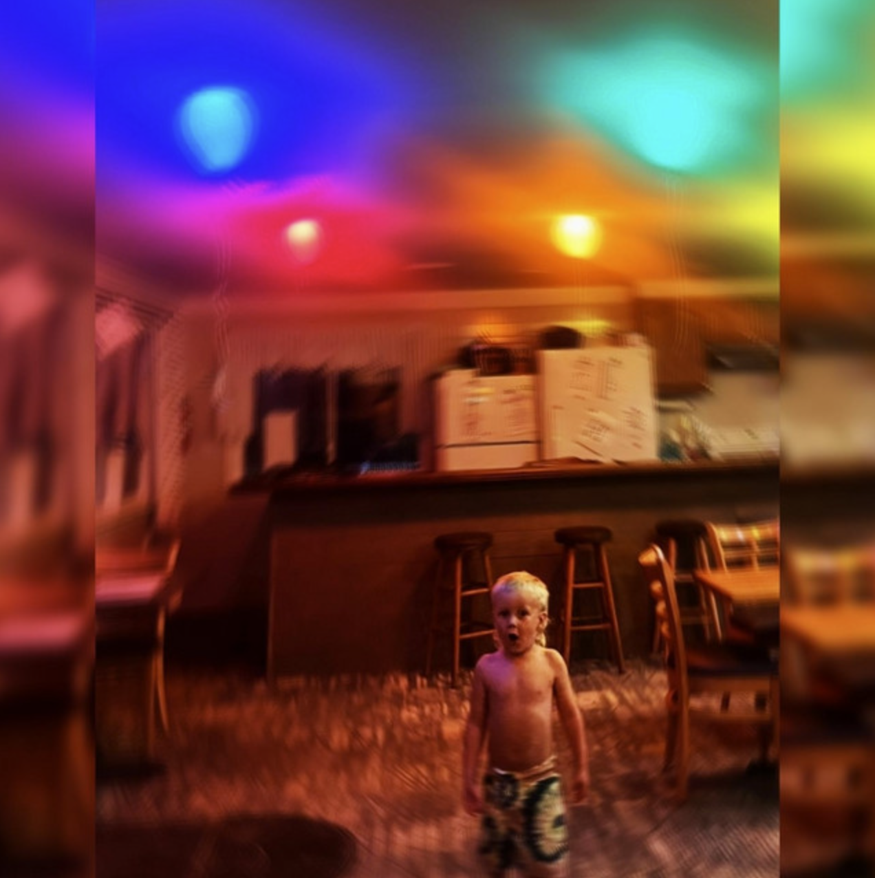 A young shirtless boy with blonde hair and patterned shorts standing in a kitchen or bar area at night, illuminated by colorful, blurred neon or party lights in pink, blue, green, and yellow.