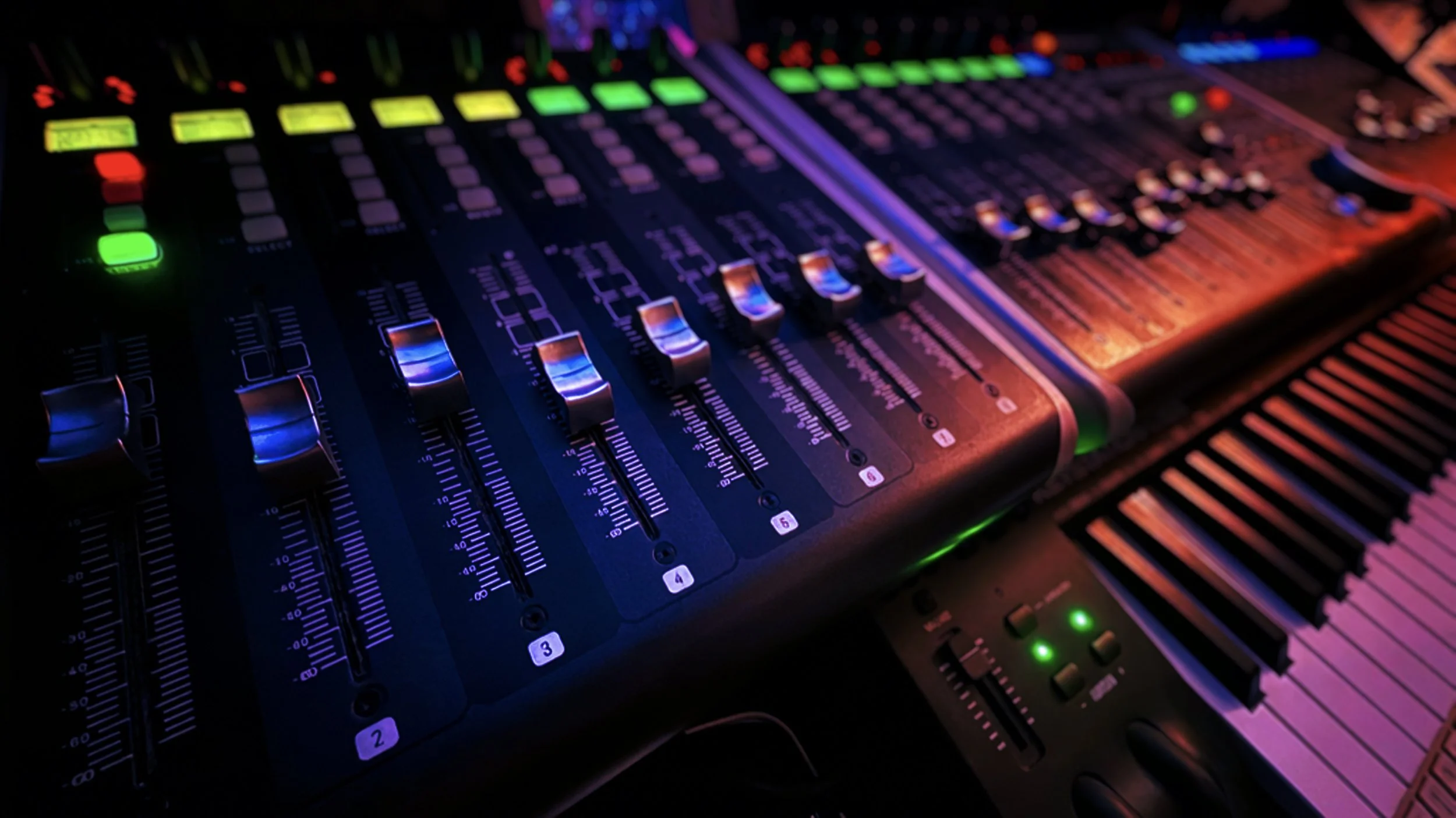 A close-up of an electronic music mixing console illuminated with colorful LED lights, showing various sliders, knobs, and controls.