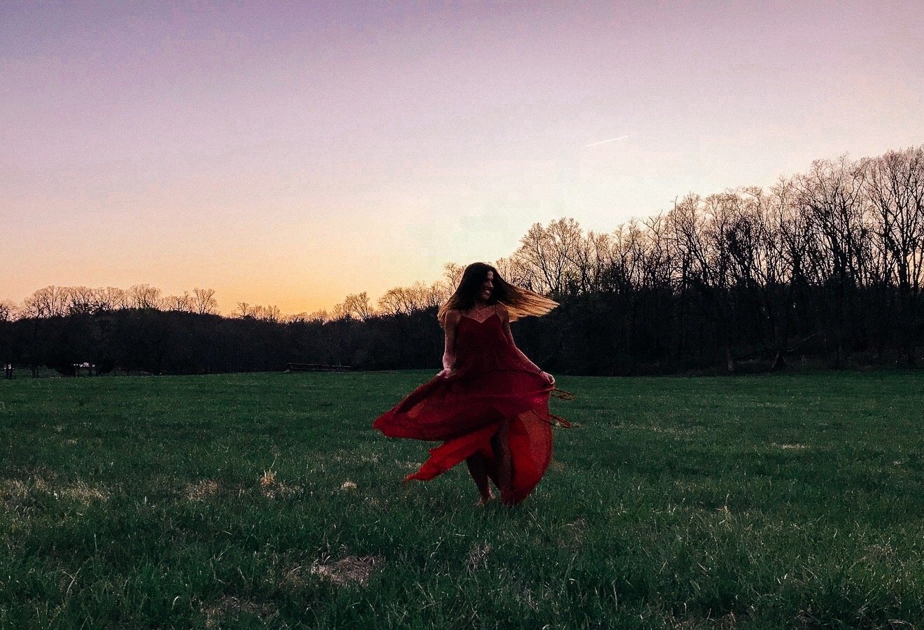 A woman in a flowing red dress dancing or spinning in an open grassy field during sunset with trees in the background.