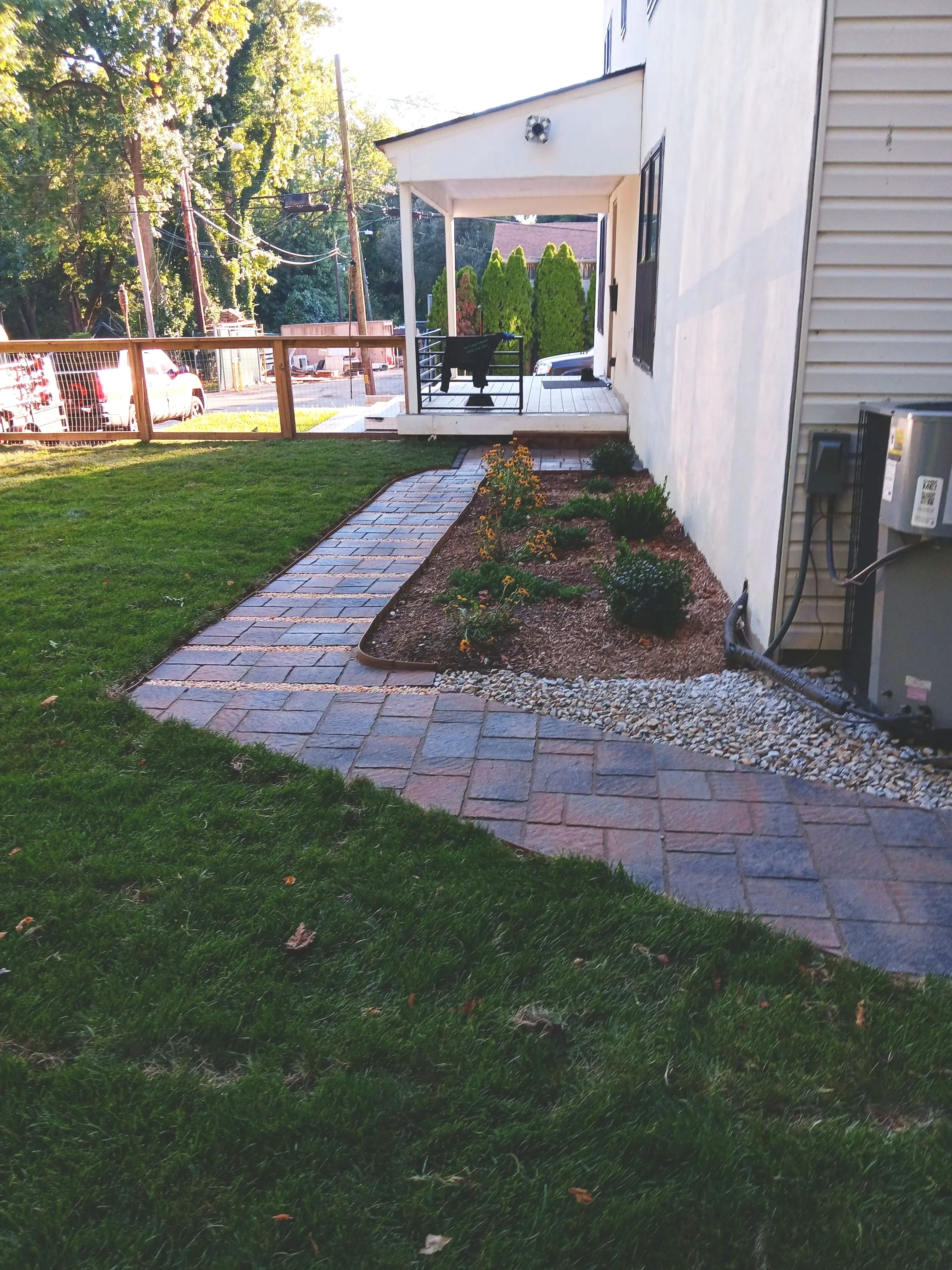 A backyard with a stone pathway leading to a porch, featuring a small garden bed with flowers and bushes, on a sunny day.