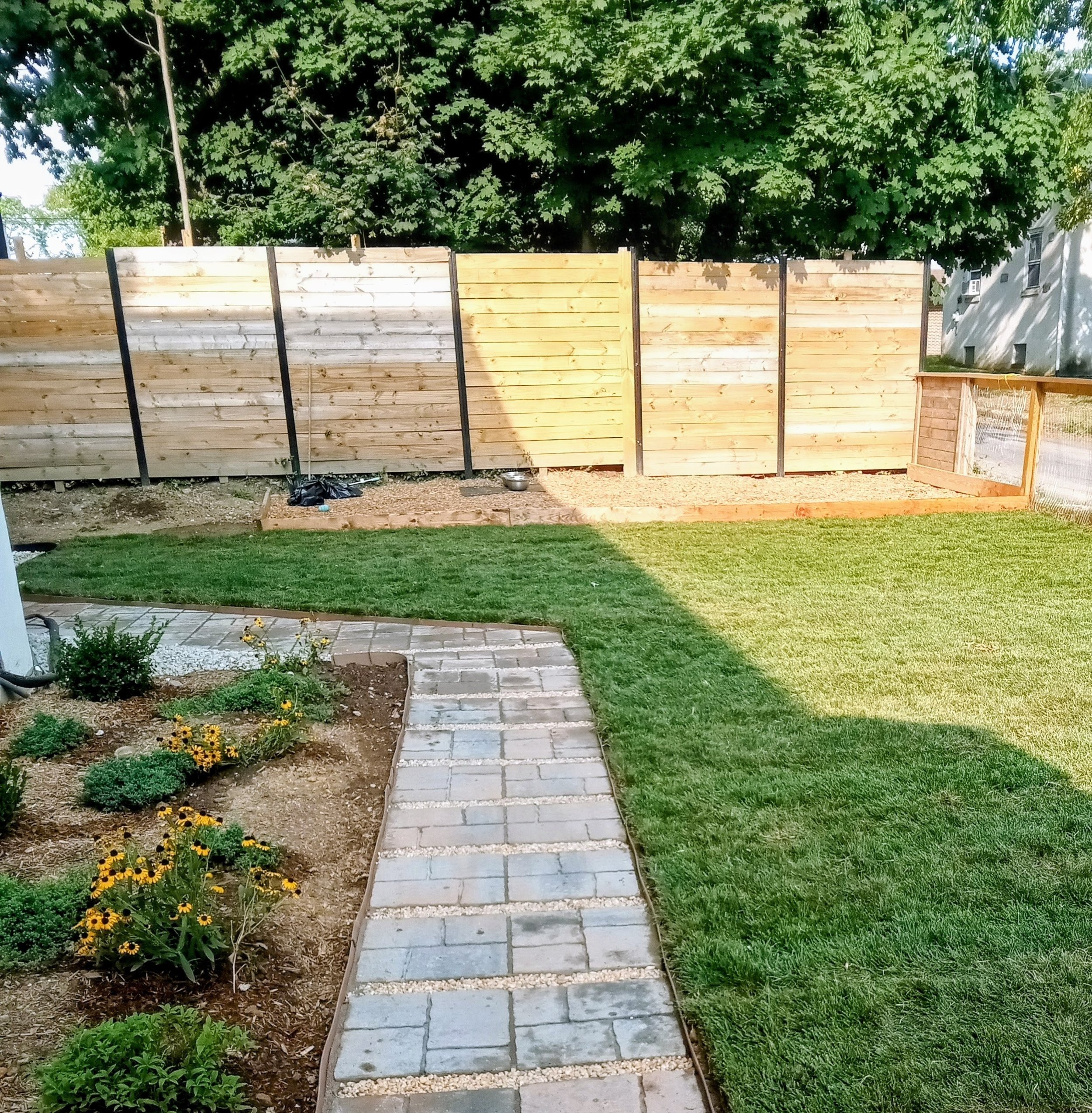 A backyard with a stone pathway, a garden bed with flowers, a well-maintained lawn, and a wooden privacy fence under a large leafy tree.