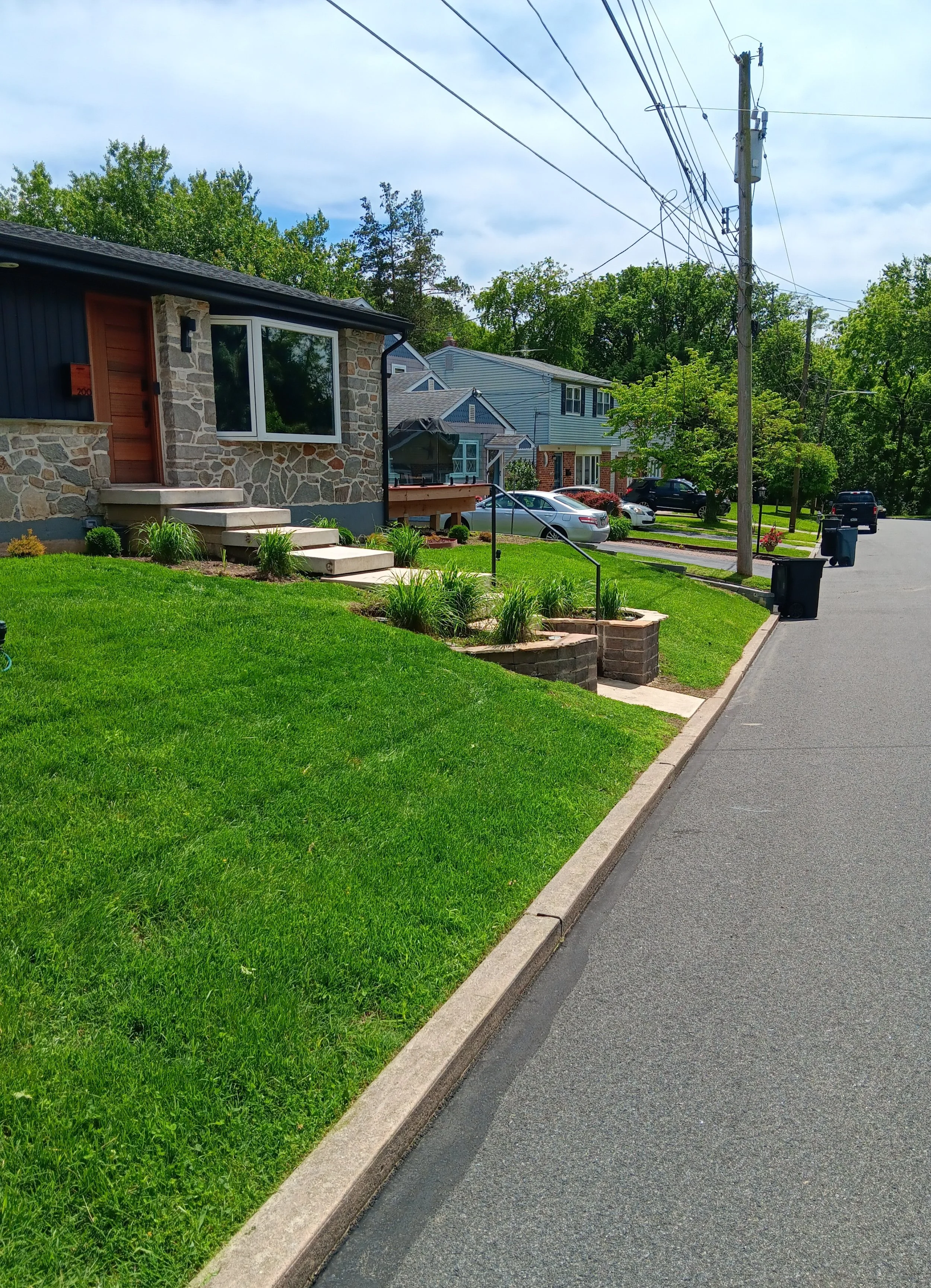 A residential neighborhood street with well-maintained lawns, houses with stone and siding exteriors, parked cars, and overhead power lines on a sunny day with partly cloudy sky.