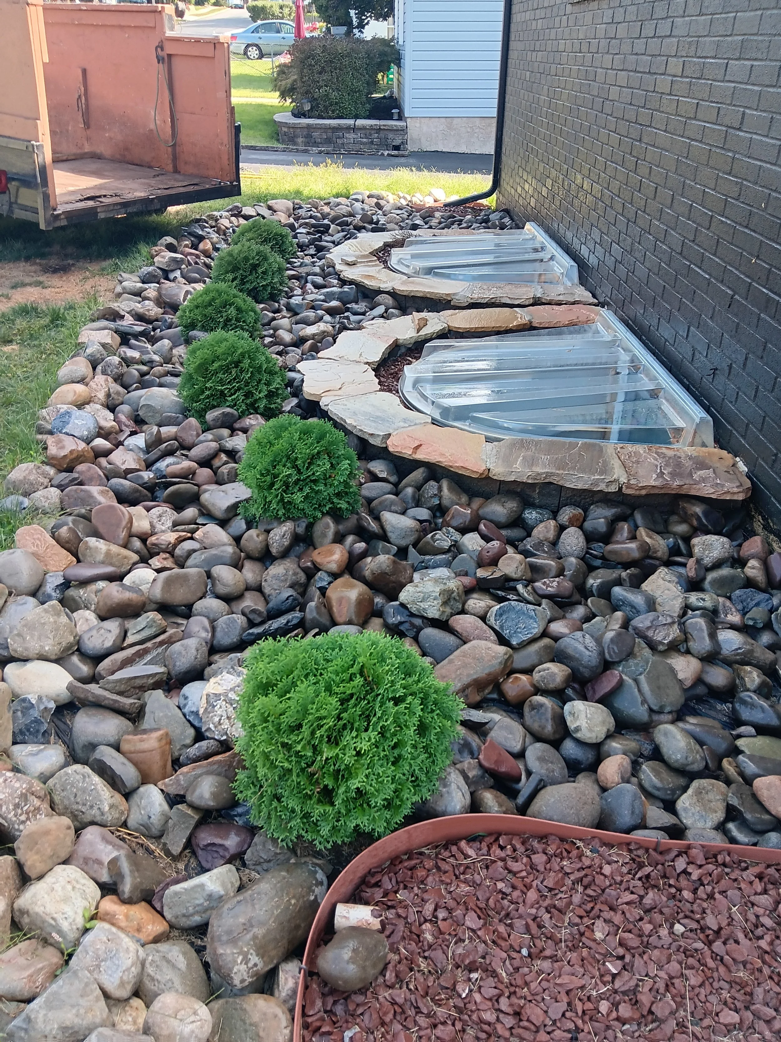 A backyard landscape with a rock garden, small green bushes, and three wooden framed glass skylights installed near a brick house wall. There is a trailer on the left side and a lawn area with trees and a white building in the background.