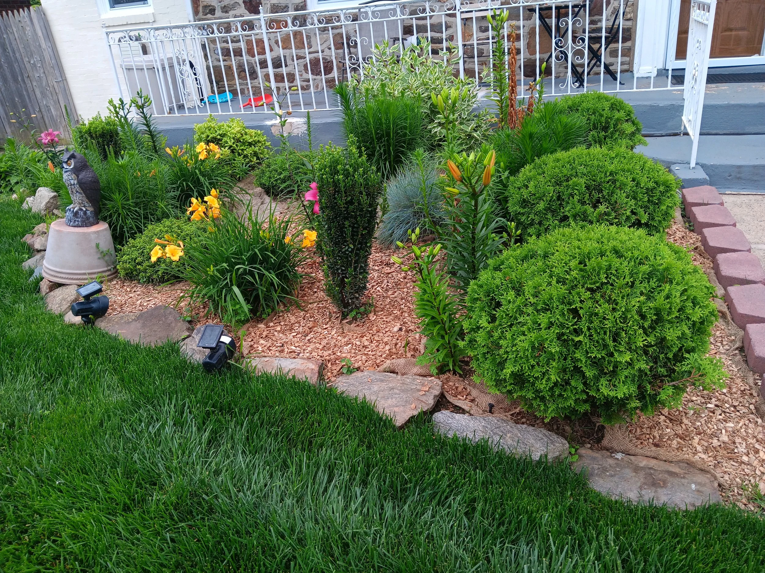 Front yard garden with various green bushes, yellow and pink flowers, decorative owl sculpture on a pedestal, solar lights, and a brick pathway near steps leading to a porch.