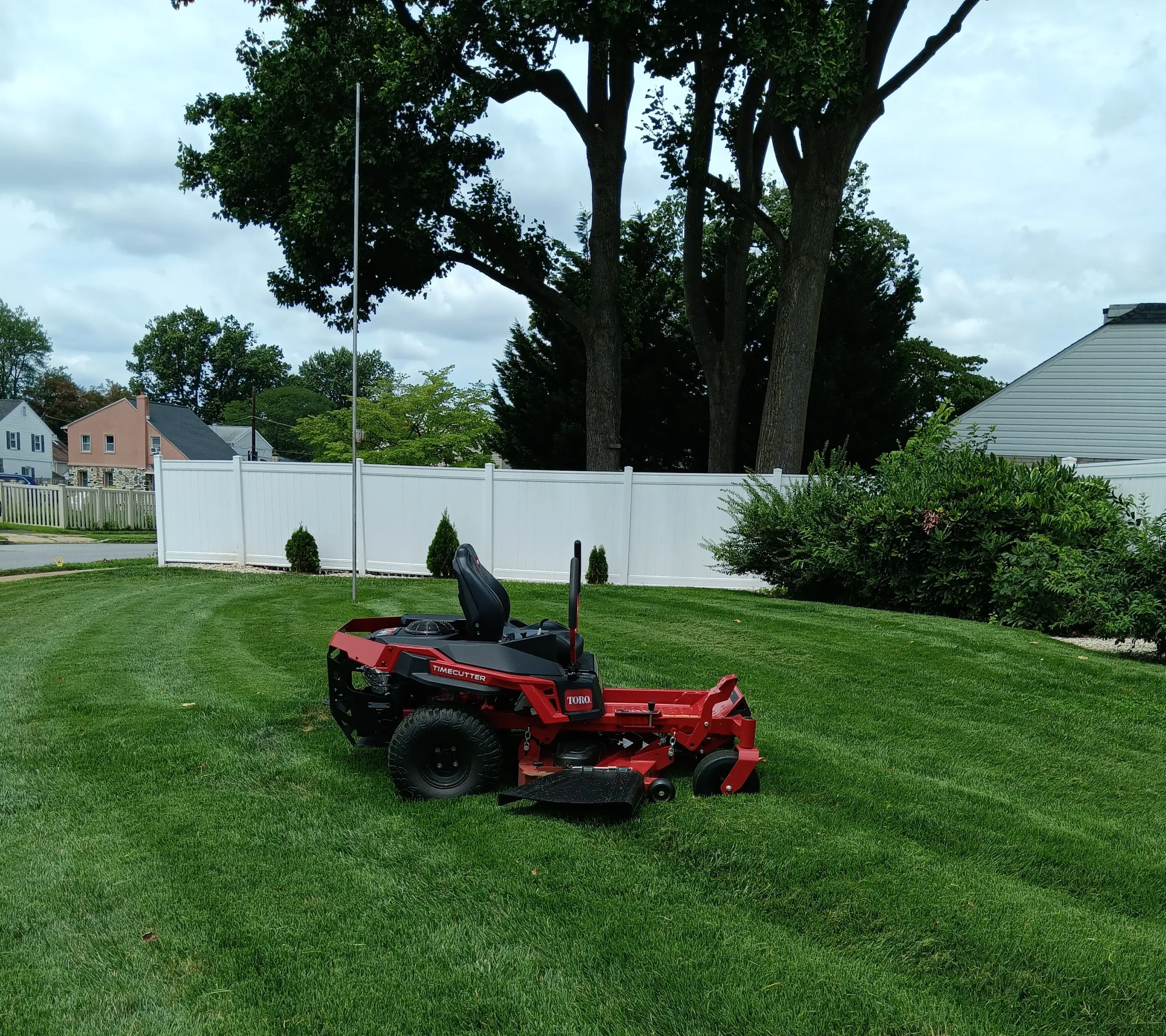 Red zero-turn lawn mower on a green, freshly mowed lawn, with a white fence, trees, and houses in the background.