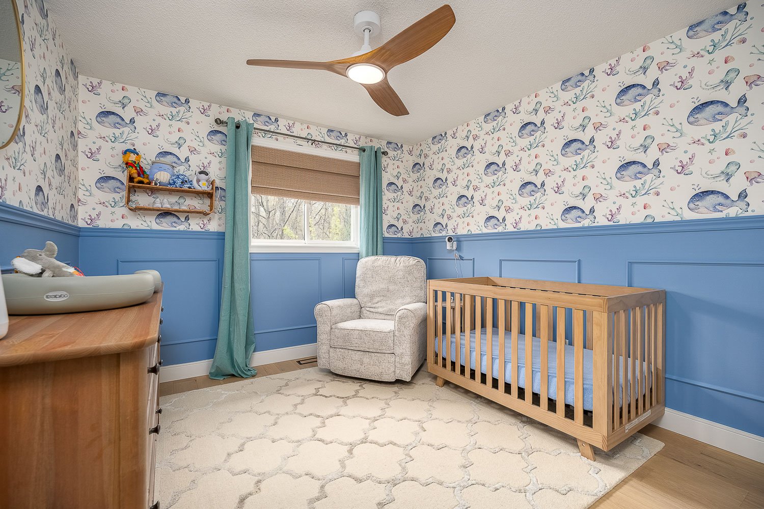 Charming nursery with custom wainscoting, whimsical ocean-inspired wallpaper, abundant natural light, and a cozy layout designed for comfort and relaxation.
