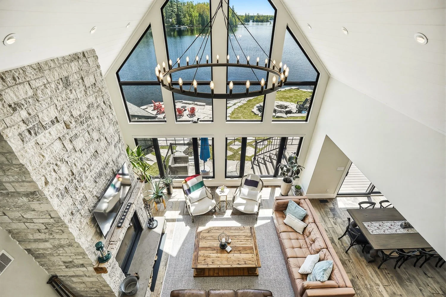 Dramatic great room captured from above, showcasing soaring cathedral ceilings, a floor-to-ceiling stone fireplace, panoramic waterfront views, and an open-concept design filled with natural light