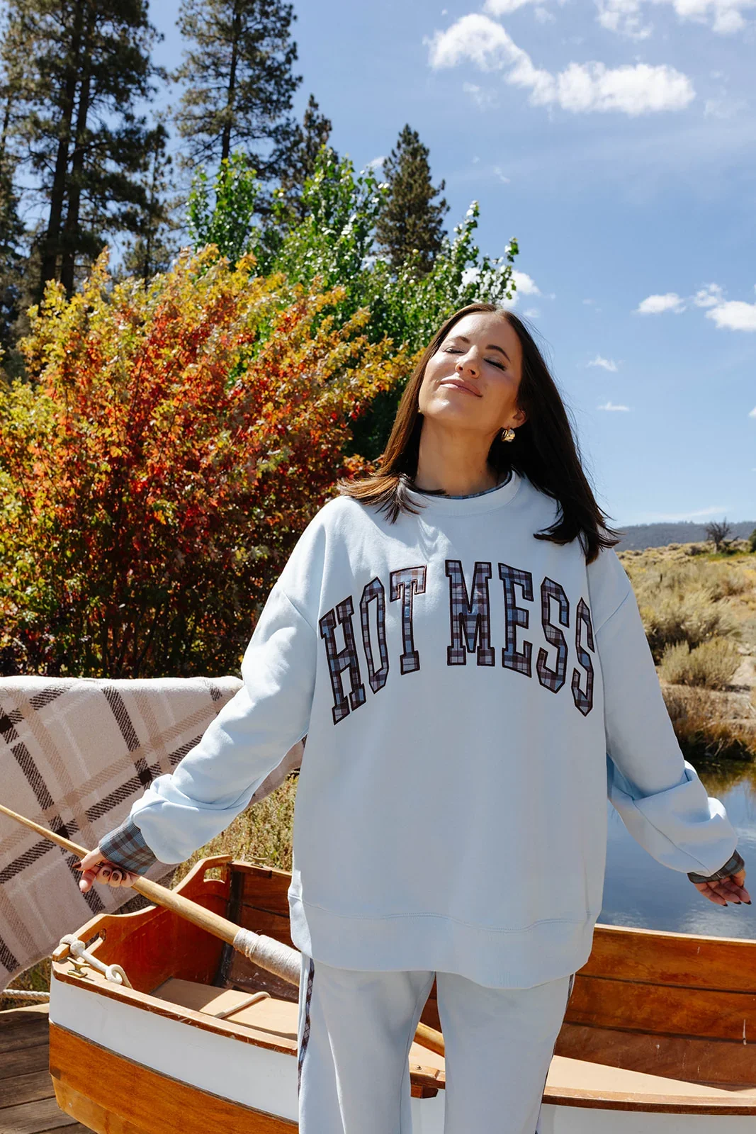  Krista standing in a scenic sunny forest, wearing a light blue sweat set with plaid writing “Hot mess” 
