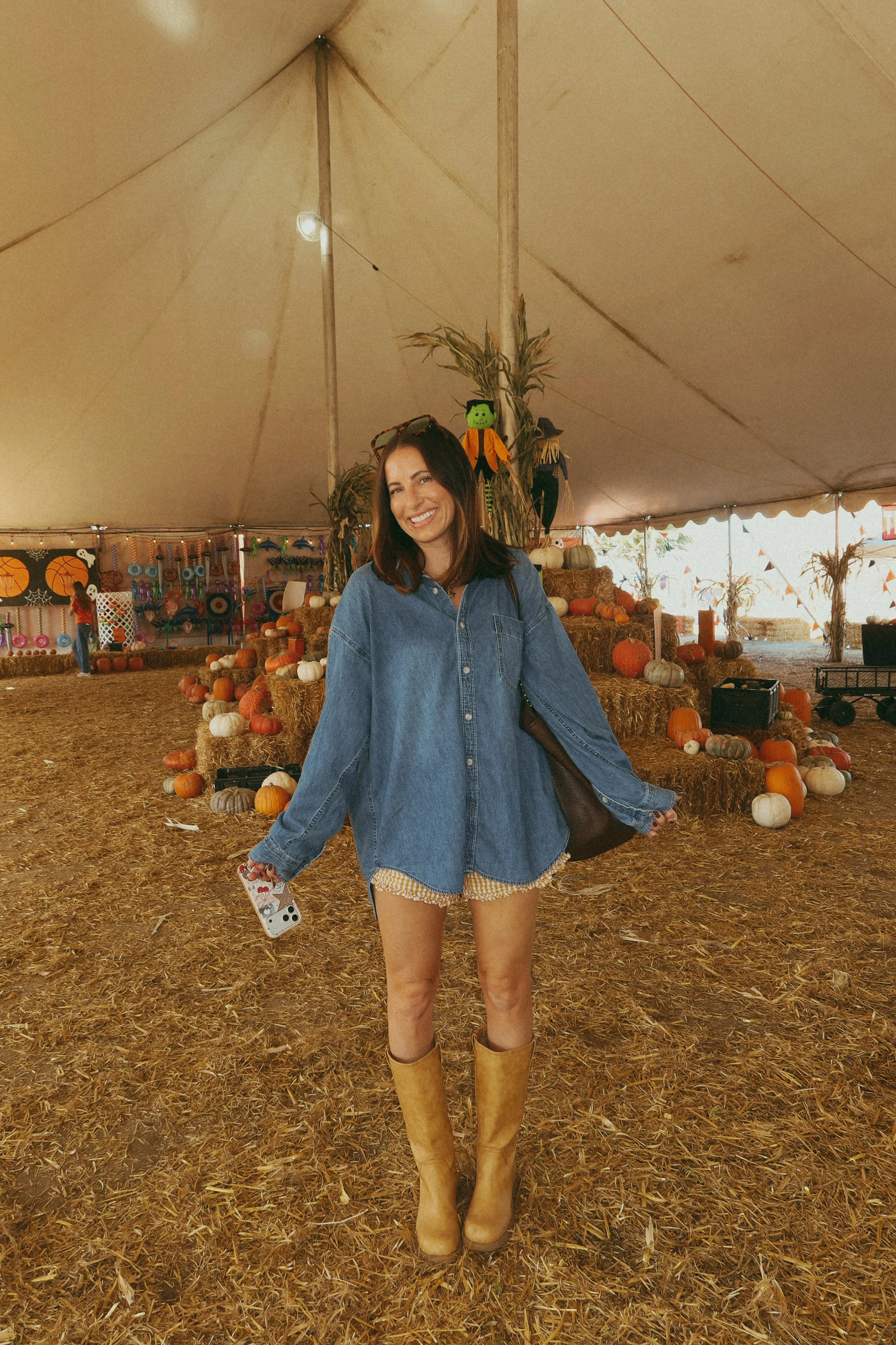 krista posing at a pumpkin patch in a fall inspired outfit. Oversized denim top, gingham printed shorts and boots. carrying her brown oversized purse with sunglasses on her head.
