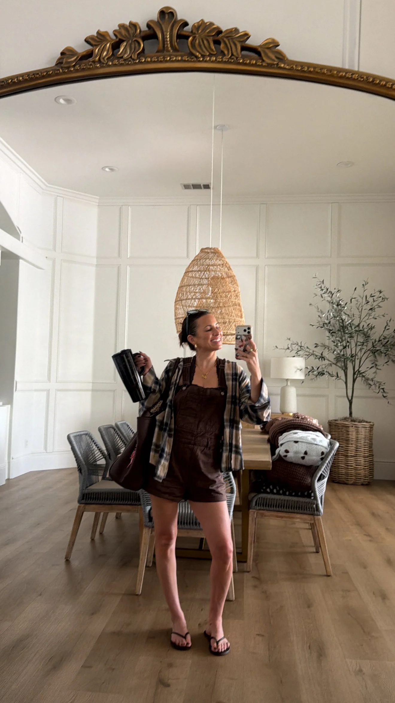 Krista posing for a mirror selfie of her outfit. Wearing brown short overalls and a plaid oversized button up over it, flip flops, hair slicked back. Also carrying a insulated brumate cup and carrying her brown shoulder bag.