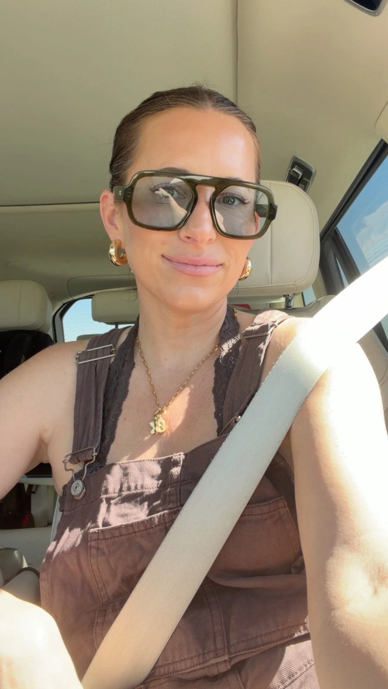 Krista taking a car selfie, wearing gold earrings, a gold necklace, oversized olive sunglasses and hair slicked back in a bun.