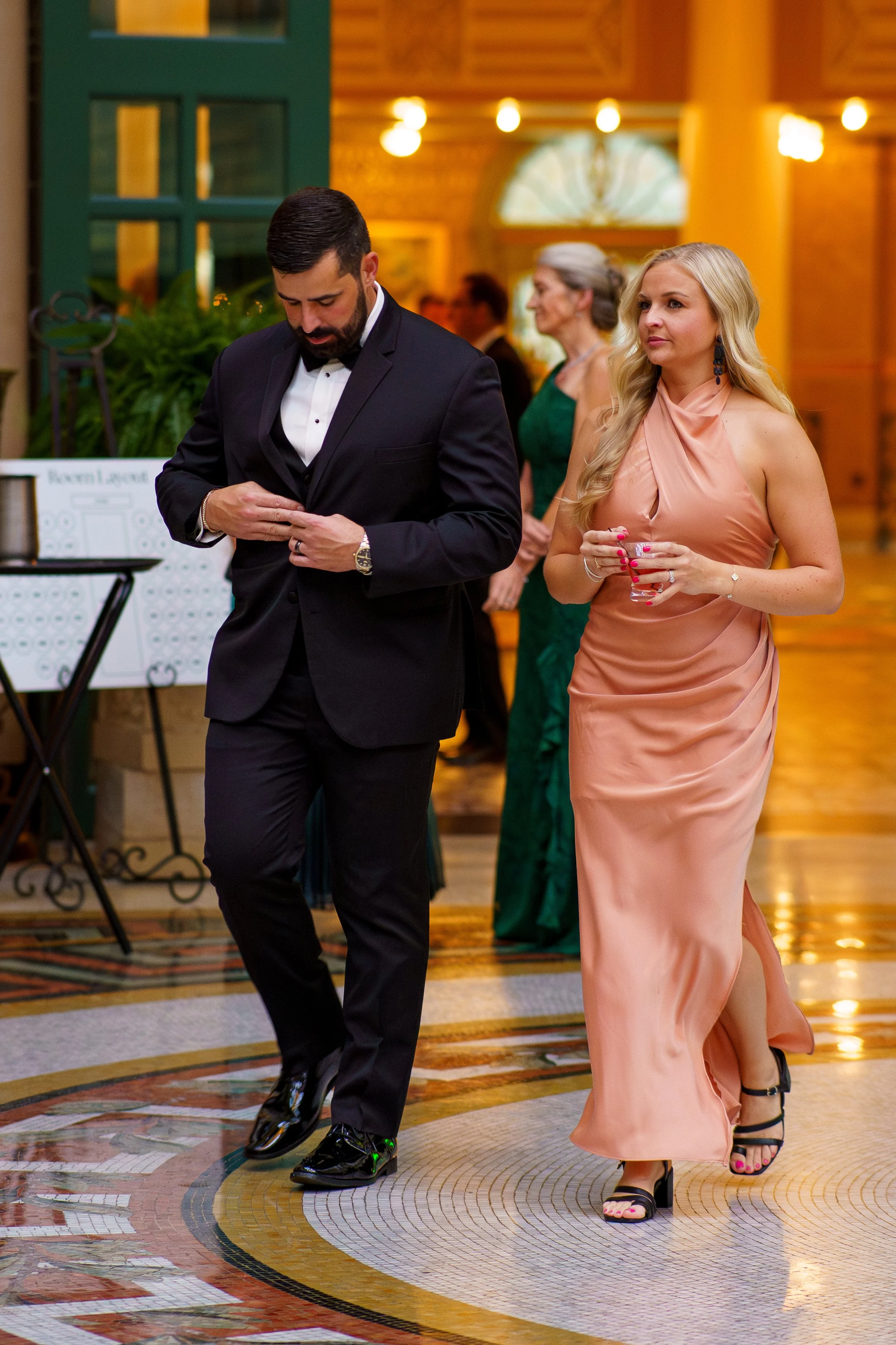 Man in a tuxedo and woman in a peach evening gown walking on a patterned floor at an event or wedding reception.