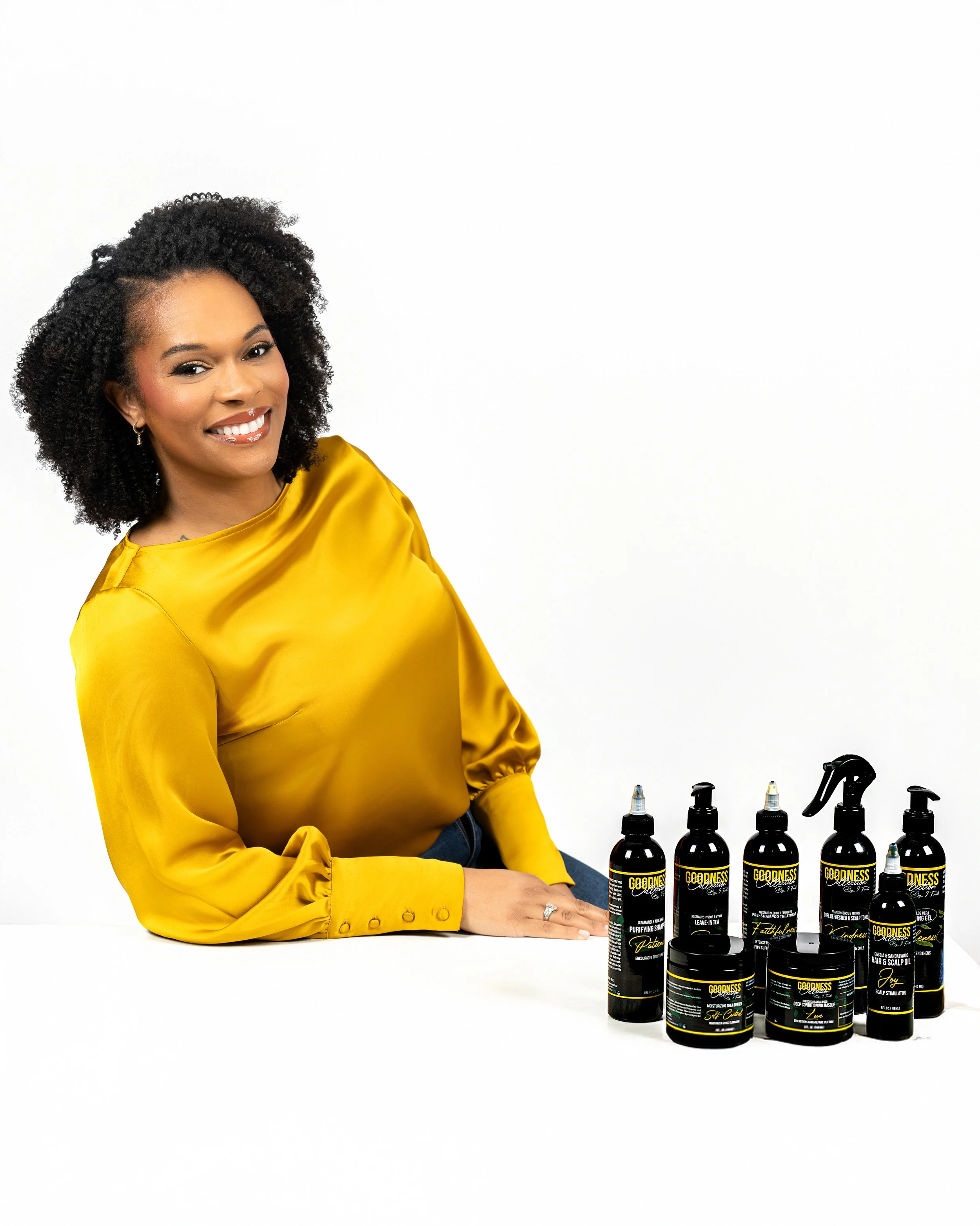 A woman with curly black hair wearing a yellow satin blouse, sitting at a white table with a lineup of black spray bottles and jars labeled 'GODDESS' for hair care products.