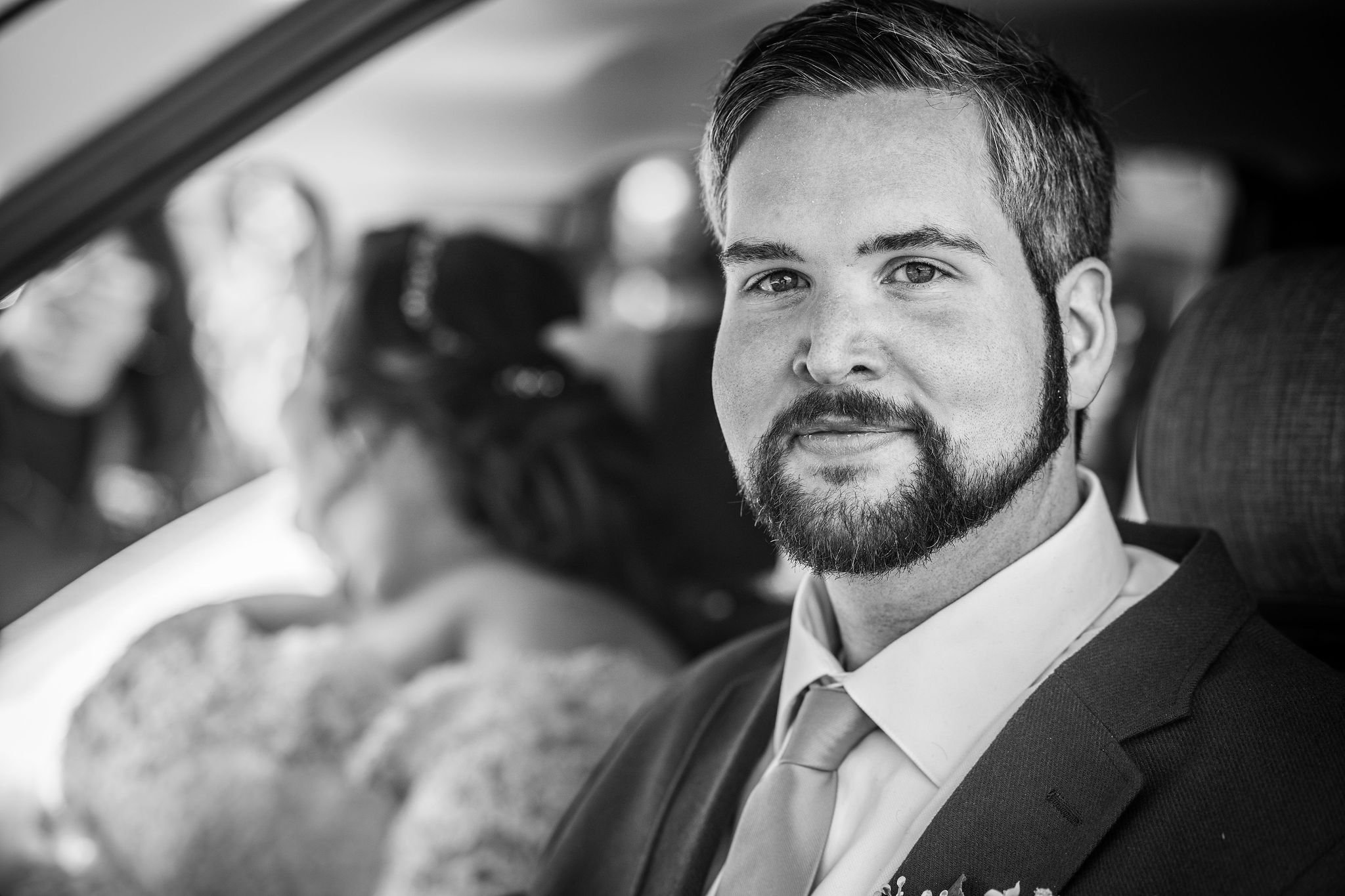 A man with a beard and suit sitting in a car, smiling at the camera. A woman with glasses and a headband is in the background.