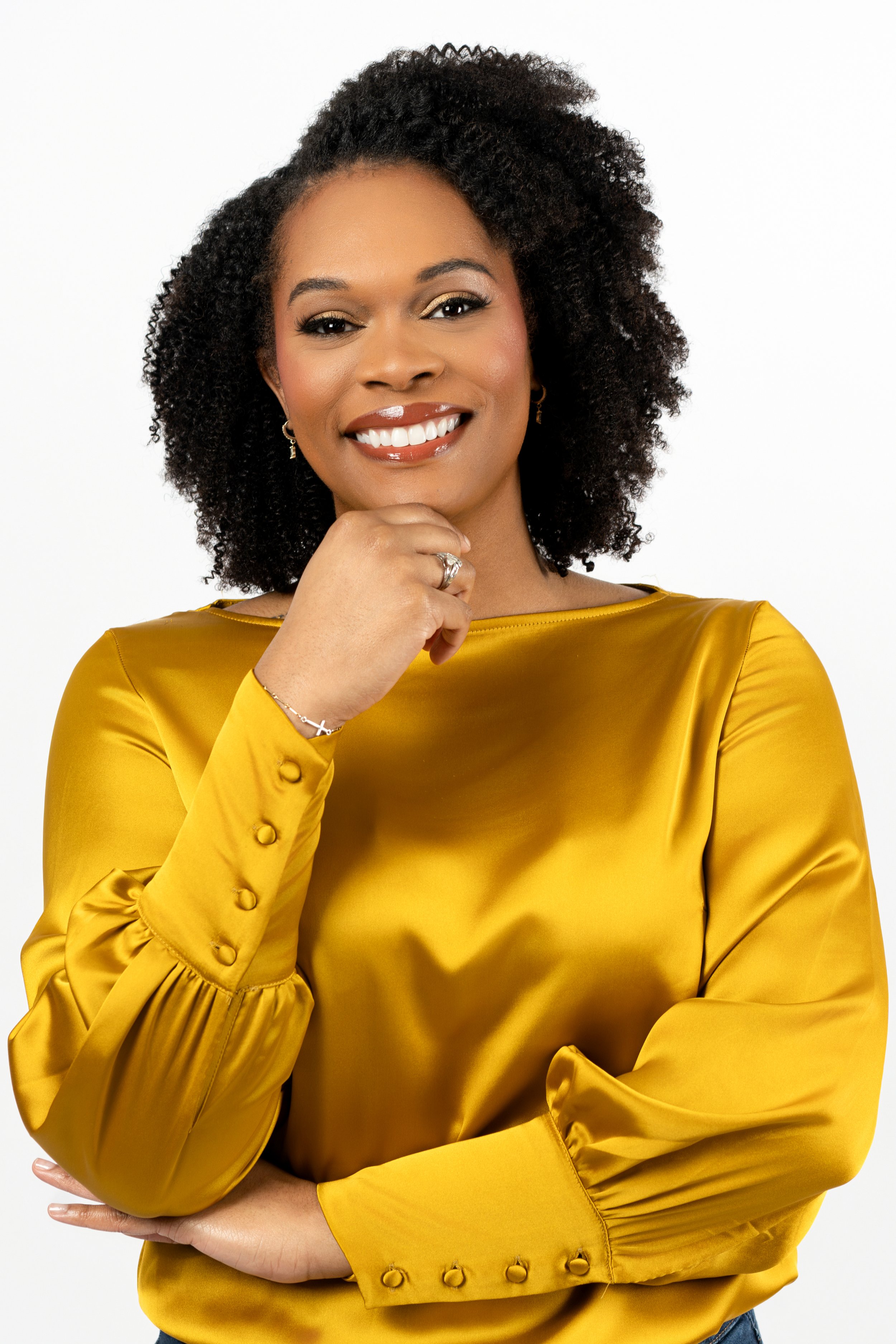 A woman with curly black hair, smiling, wearing a gold satin long-sleeve blouse with button details, posing with her hand near her chin against a plain white background.