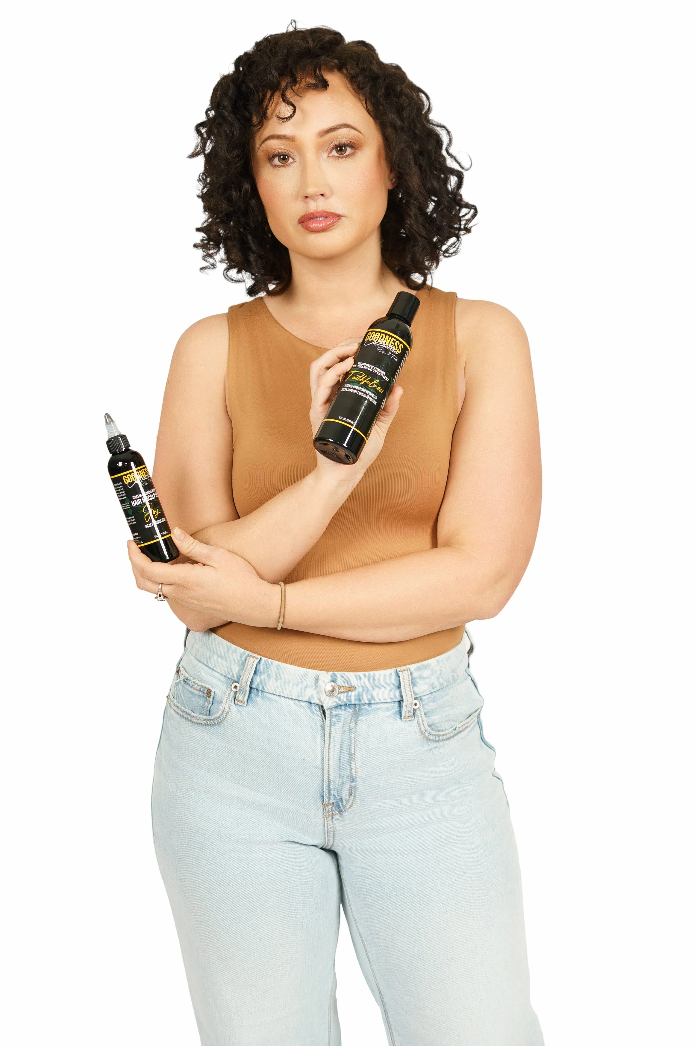A woman with curly dark hair and nude lipstick holding two bottles of hair oil against a white background.
