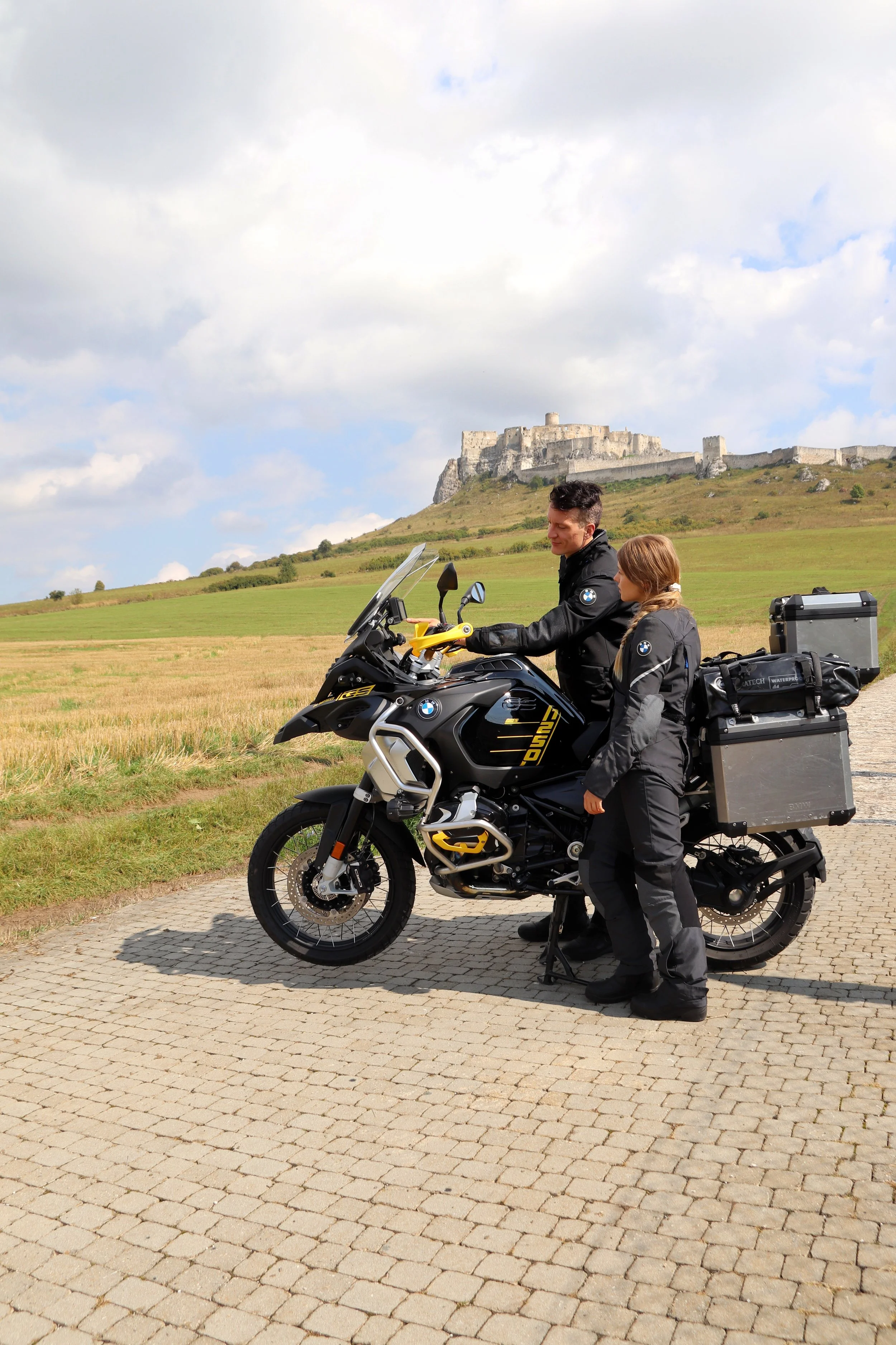 Two people in motorcycle gear standing beside a BMW adventure motorcycle on a paved path, with a hill and large stone fortress in the background.