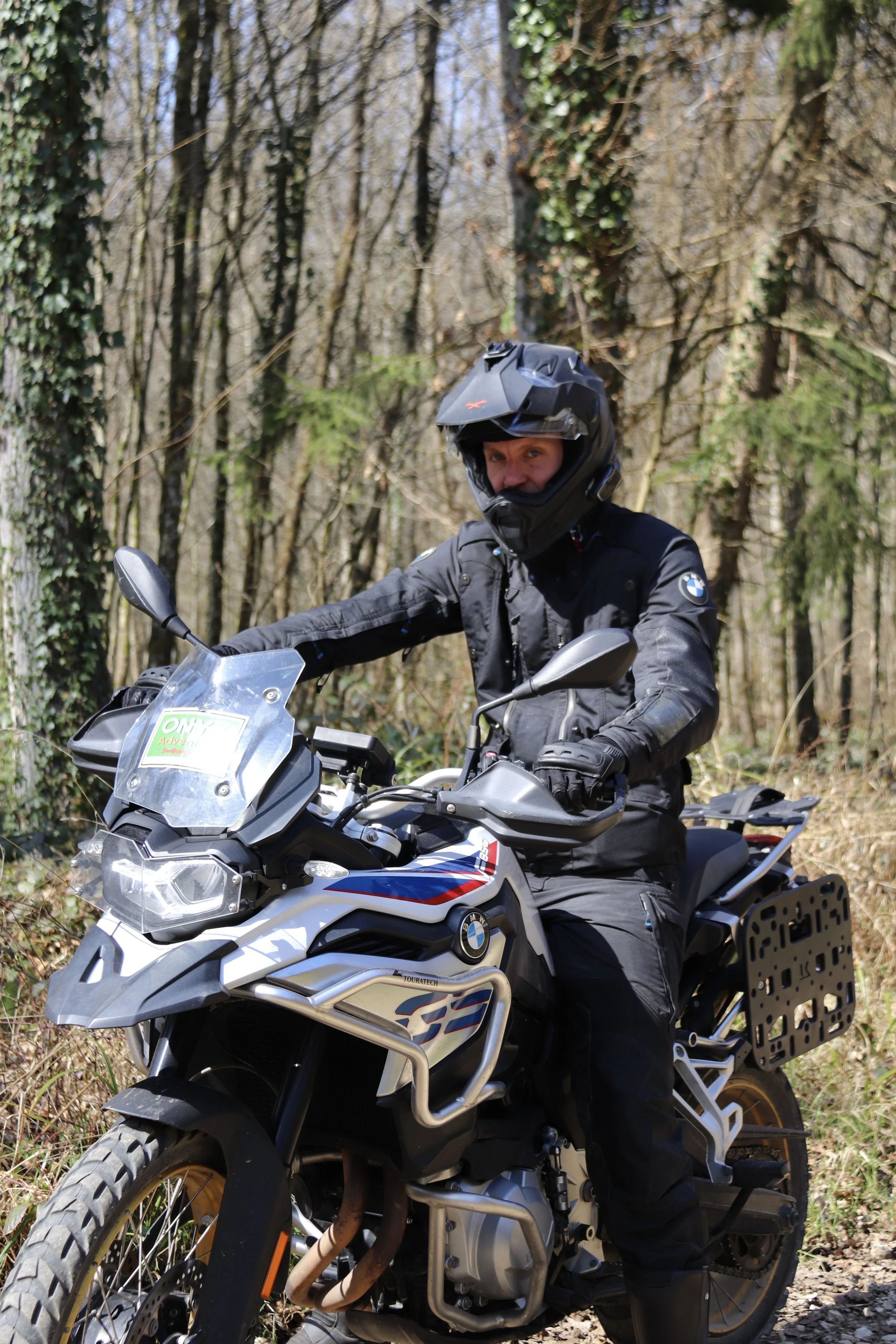 Person wearing black motorcycle gear and helmet riding a BMW adventure motorcycle in a forest setting.