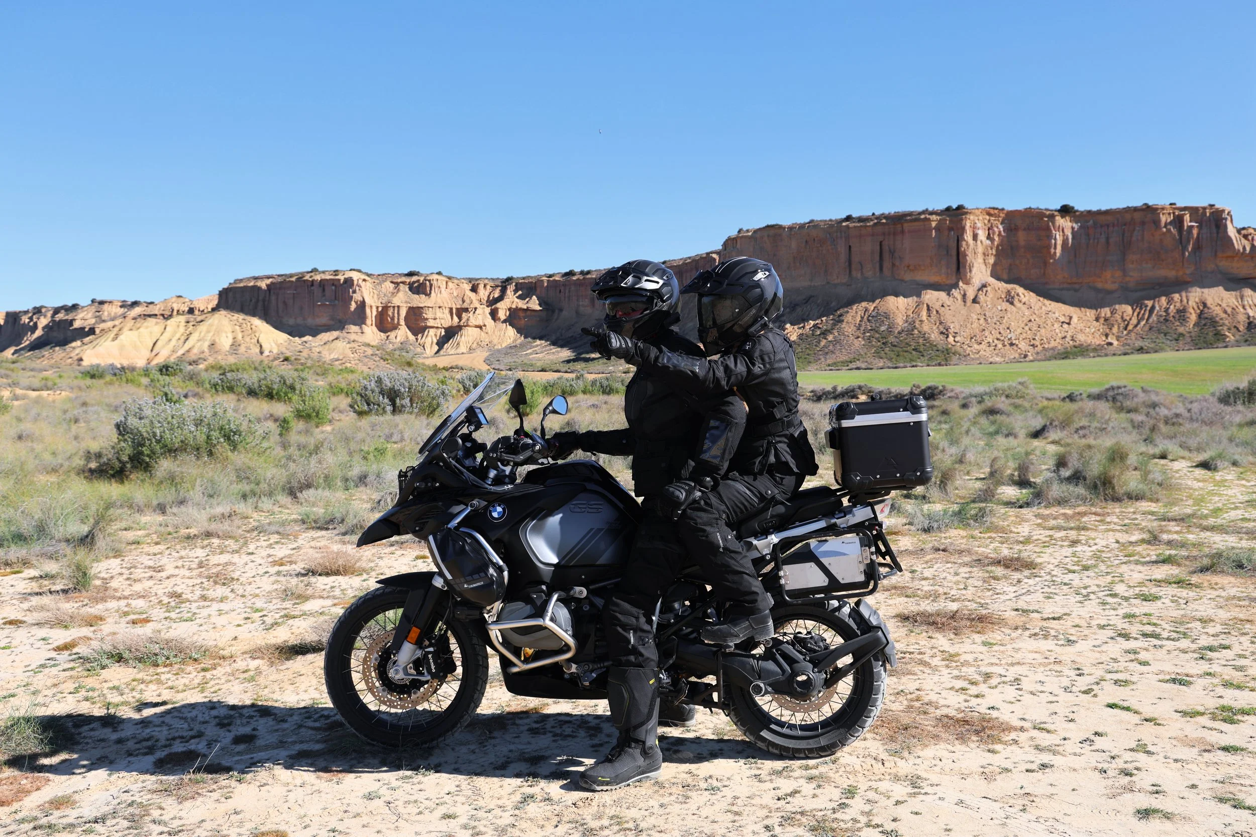 Two motorcyclists wearing black gear and helmets on an adventure motorcycle in a desert landscape with mesas and clear blue sky.