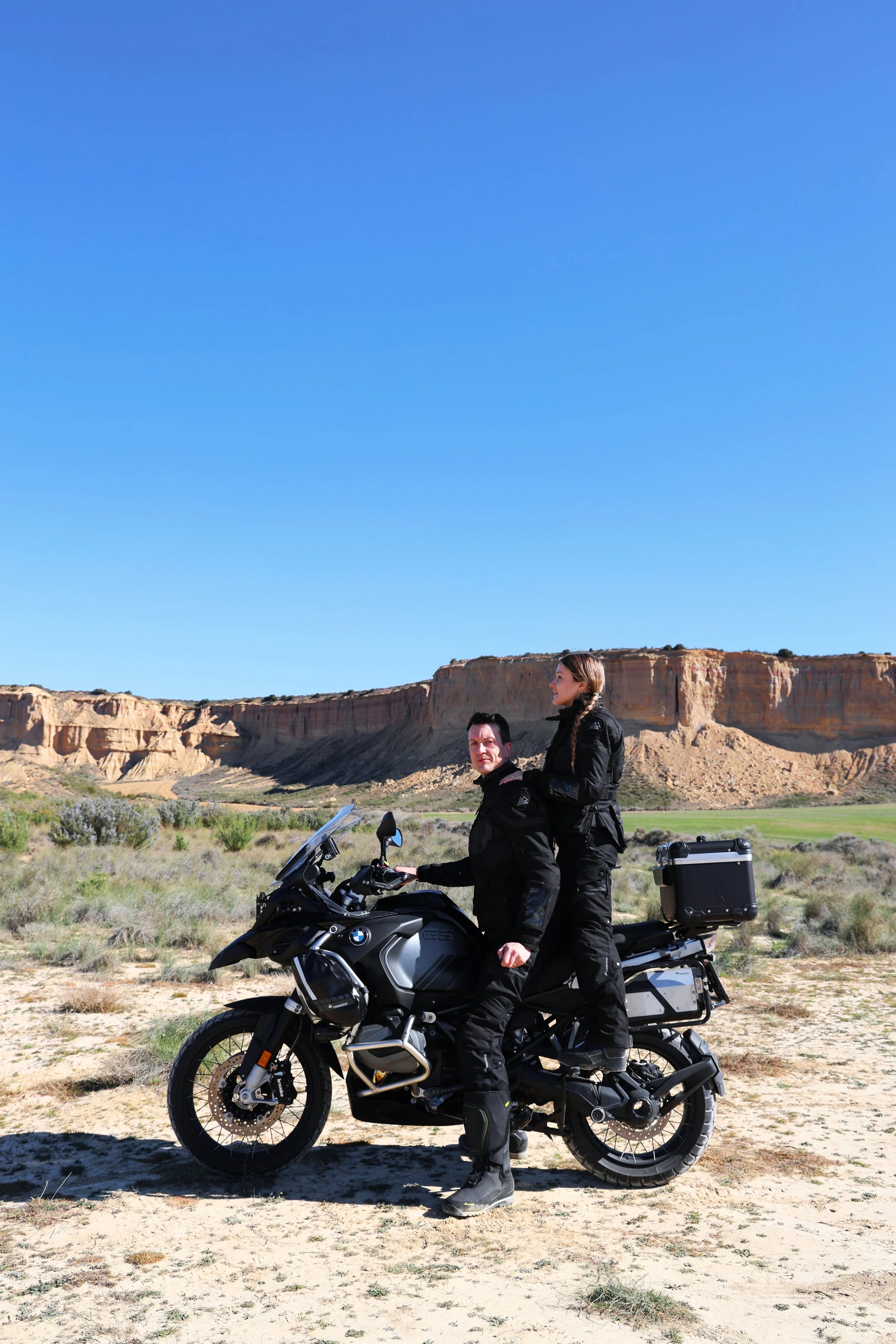 Two people on a black motorcycle in a desert landscape with tall rock formations under a clear blue sky.