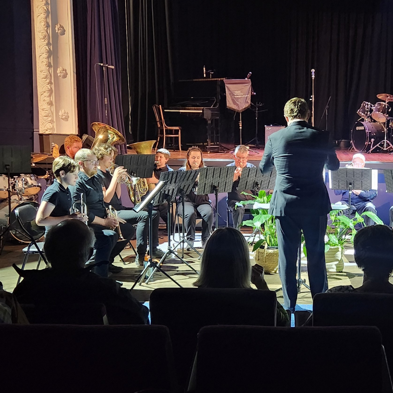 Image of a YRC orchestra conductor standing in front of a band of younger and older people playing brass instruments