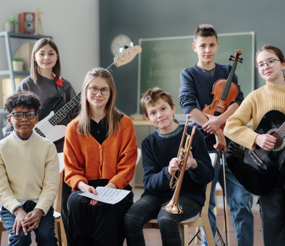 a group of young people with various instruments sitting together and smiling to camera. They are holding a guitar, sheet music, violin, trumpet and a bass guitar
