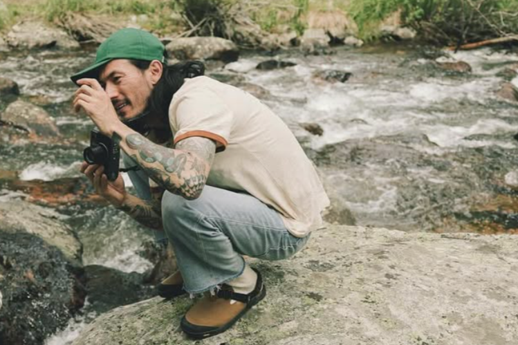 Person with tattoos and a green cap crouching on rocks by a stream, holding a camera.