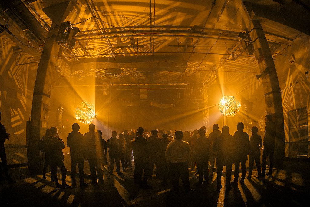 A crowd of people gathered in a dark indoor space illuminated by warm yellow-orange lights, with shadows and patterns cast on the walls and ceiling.