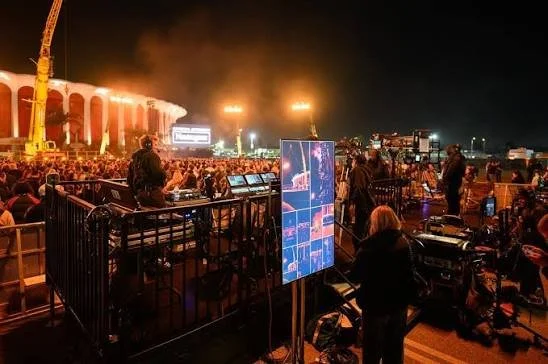Crowd of people at an outdoor event at night, with a stage or structure illuminated in the background and equipment set up for broadcasting or recording.