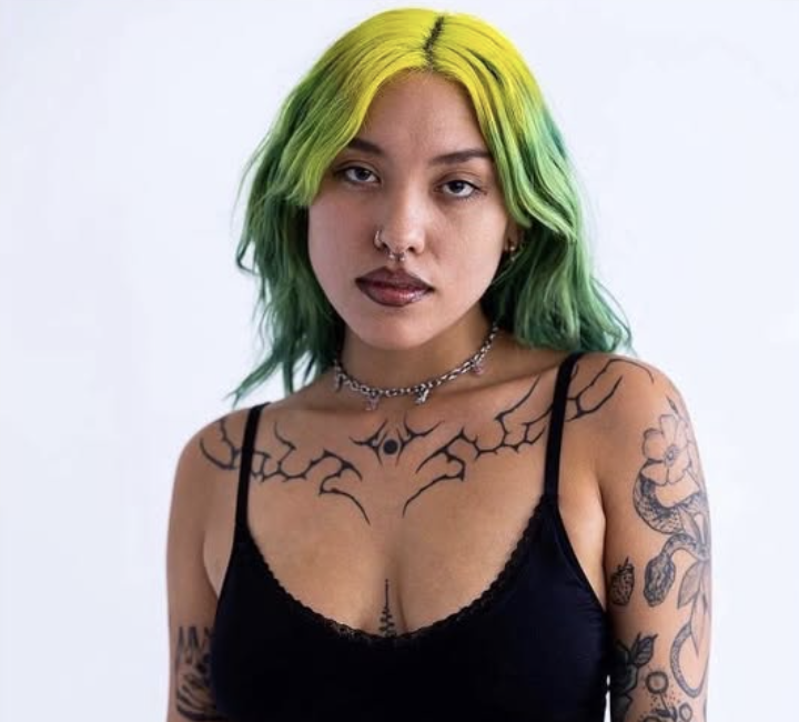 A young woman with green and yellow hair, wearing a black tank top, tattoos on her arms and chest, a septum piercing, and a choker necklace, against a plain white background.